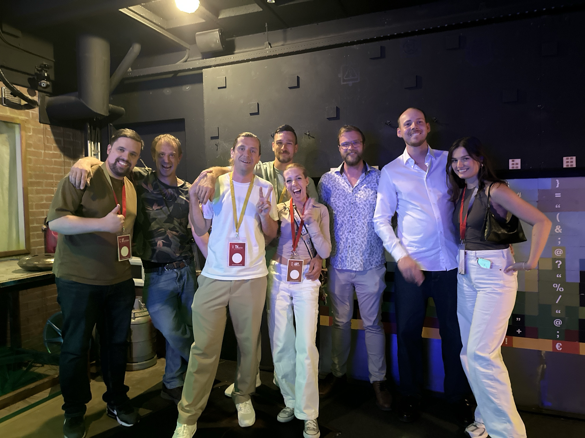 The image shows a group of eight people, six men and two women, posing for a photo indoors. The background appears to be a dark, textured wall with some decorative elements, possibly in a venue or studio. The lighting is dim, with overhead lights providing illumination. The people are casually dressed, with some wearing lanyards and identification tags, suggesting they might be at an event or conference. They are standing close together, with most of them smiling and looking at the camera. One woman in the center is making a peace sign with her fingers and appears to be laughing. Another woman on the right is posing with her hand on her hip. The overall atmosphere seems friendly and relaxed. There are no specific visual cues in the image that would definitively place it in Amsterdam, Netherlands.