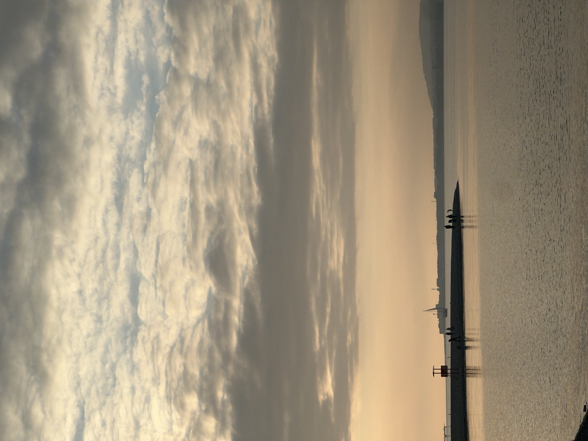 The image displays a coastal scene in Gelendzhik, Russia, under soft, warm light indicative of sunrise or sunset. The upper two-thirds of the frame are occupied by a cloudy sky, featuring layered altocumulus and stratus formations illuminated with golden-grey and white tones, transitioning to darker grey overhead. The lower third shows a calm body of water, likely the Black Sea, reflecting the sky's warm hues with minimal ripples.
A dark-surfaced pier or breakwater extends from the left into the water. A red-painted, two-tiered lifeguard stand or observation tower with a small roof is situated on the leftmost section of this structure. Further along the pier, approximately 5-7 individuals are visible as dark silhouettes, either standing or walking. A second, smaller group of 2-3 silhouetted individuals is positioned further to the right on the pier.
The distant horizon features the city skyline of Gelendzhik. Notable silhouettes include a sailboat with a prominent mast and sail, several industrial-style structures consistent with port facilities, and other urban buildings. To the right, the landmass rises into a series of low hills or mountains.