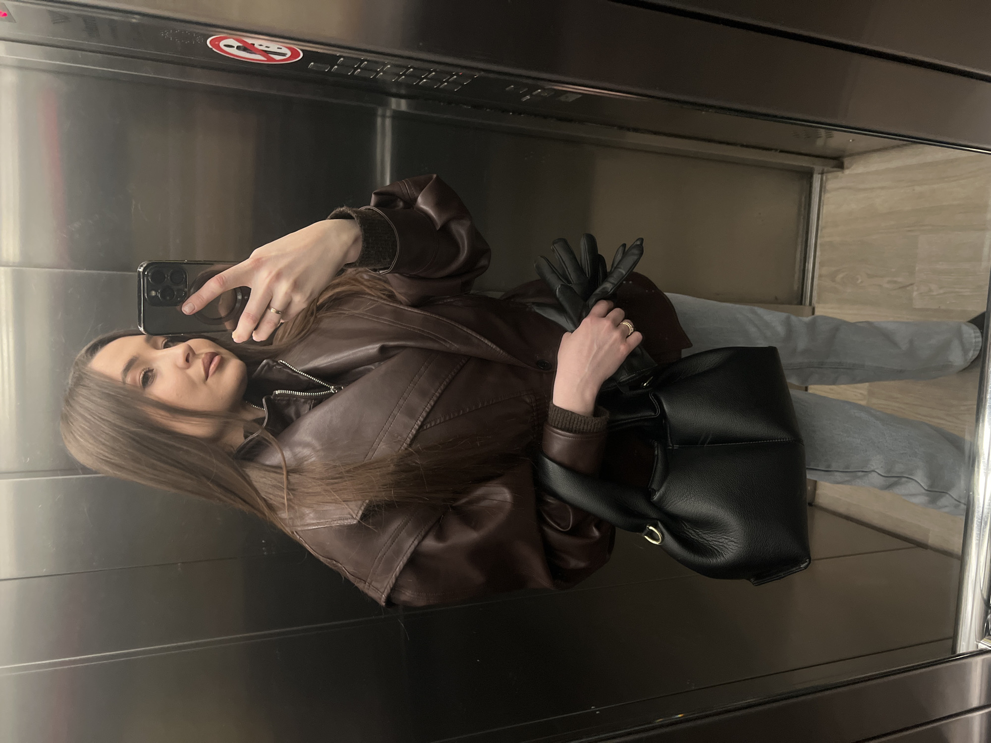 A young woman is taking a selfie in what appears to be an elevator. She is holding her phone in her right hand. The phone appears to be black and is reflecting the elevator interior. She is wearing a brown leather jacket and light blue jeans. She has long brown hair and is wearing a ring on her right hand. In her left hand, she holds a pair of black leather gloves and a black leather handbag. The elevator walls are metal and have a warning sign at the top. The elevator floor is light-colored with a tile-like pattern. The location of the image within the city of Khimki, Russia, cannot be determined.