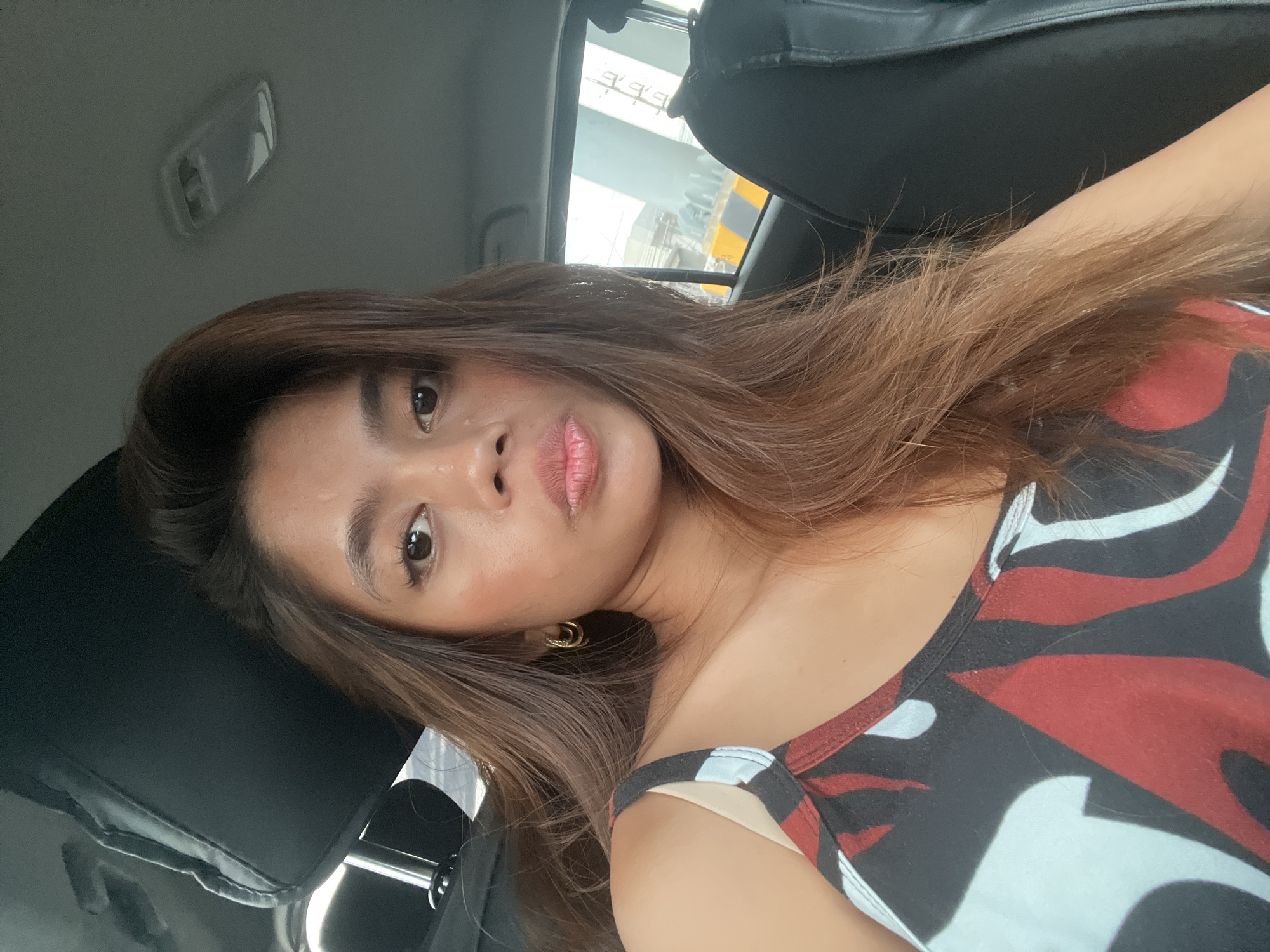 A young woman with long, brown hair is taking a selfie in a car. She is wearing a tank top with a red, black, and white abstract pattern. She has on small gold hoop earrings. The interior of the car is visible behind her, including a black headrest, the ceiling with a light fixture, and the back seat. Outside the car window, a blurred view of a bridge and some yellow and black barriers can be seen, suggesting an urban or possibly highway setting. The lighting indicates it is daytime. No specific landmarks identifying Panabo, Philippines are visible.