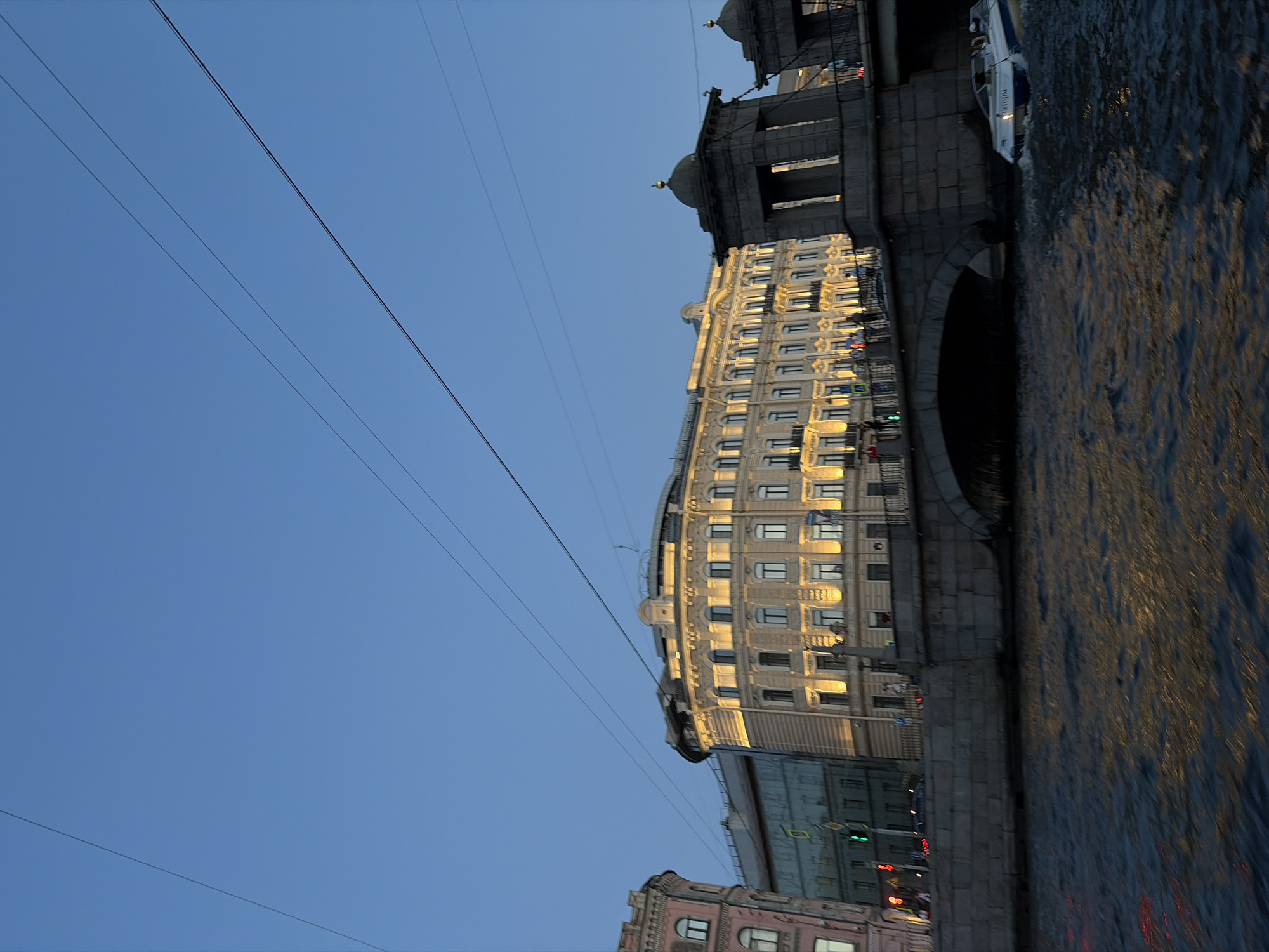 The image shows a view of the Anichkov Bridge over the Fontanka River in Saint Petersburg, Russia. The bridge is made of stone and has decorative elements. On the embankment, there is a multi-story building with a light-colored facade, illuminated by warm lights. The building has many windows and balconies. A boat is visible in the river, moving away from the viewer and towards the bridge. The sky is clear and blue, with some power lines crossing it. The water in the river reflects the lights from the buildings and the sky. The overall scene depicts a picturesque urban landscape at dusk.
