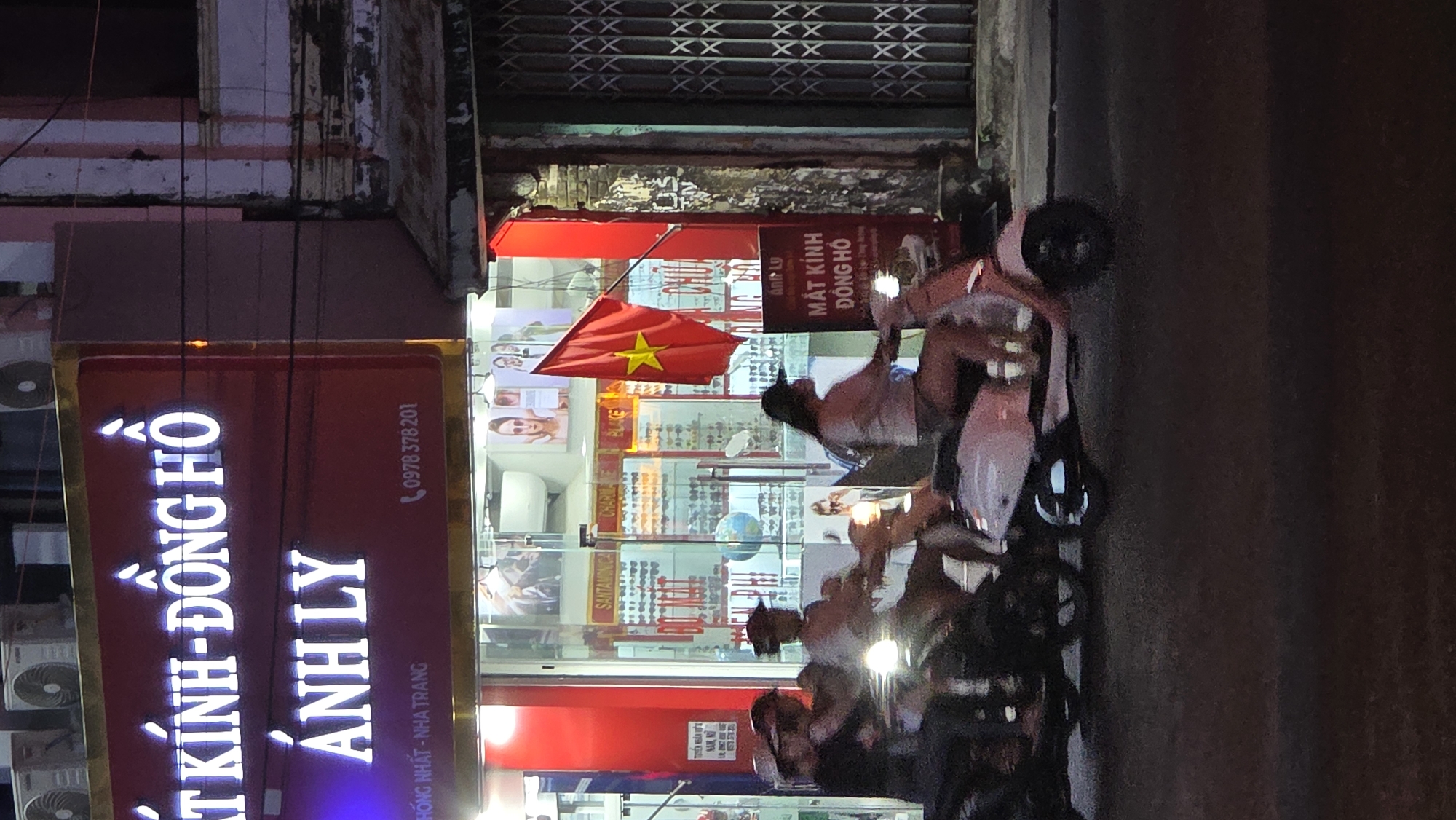 The scene depicts a street in Nha Trang, Vietnam, at night. An optics shop, "Ánh Ly Mắt Kính-Đồng Hồ" (Ánh Ly Eyewear-Watches), is prominently displayed. Its illuminated signage includes the phone number "0978 378 201". The name is written in large, illuminated Vietnamese script. A Vietnamese flag is positioned near the shop entrance. The store's interior displays various eyewear brands, including "Santamónica" and "Ánh Ly". Outside, several people are riding scooters.