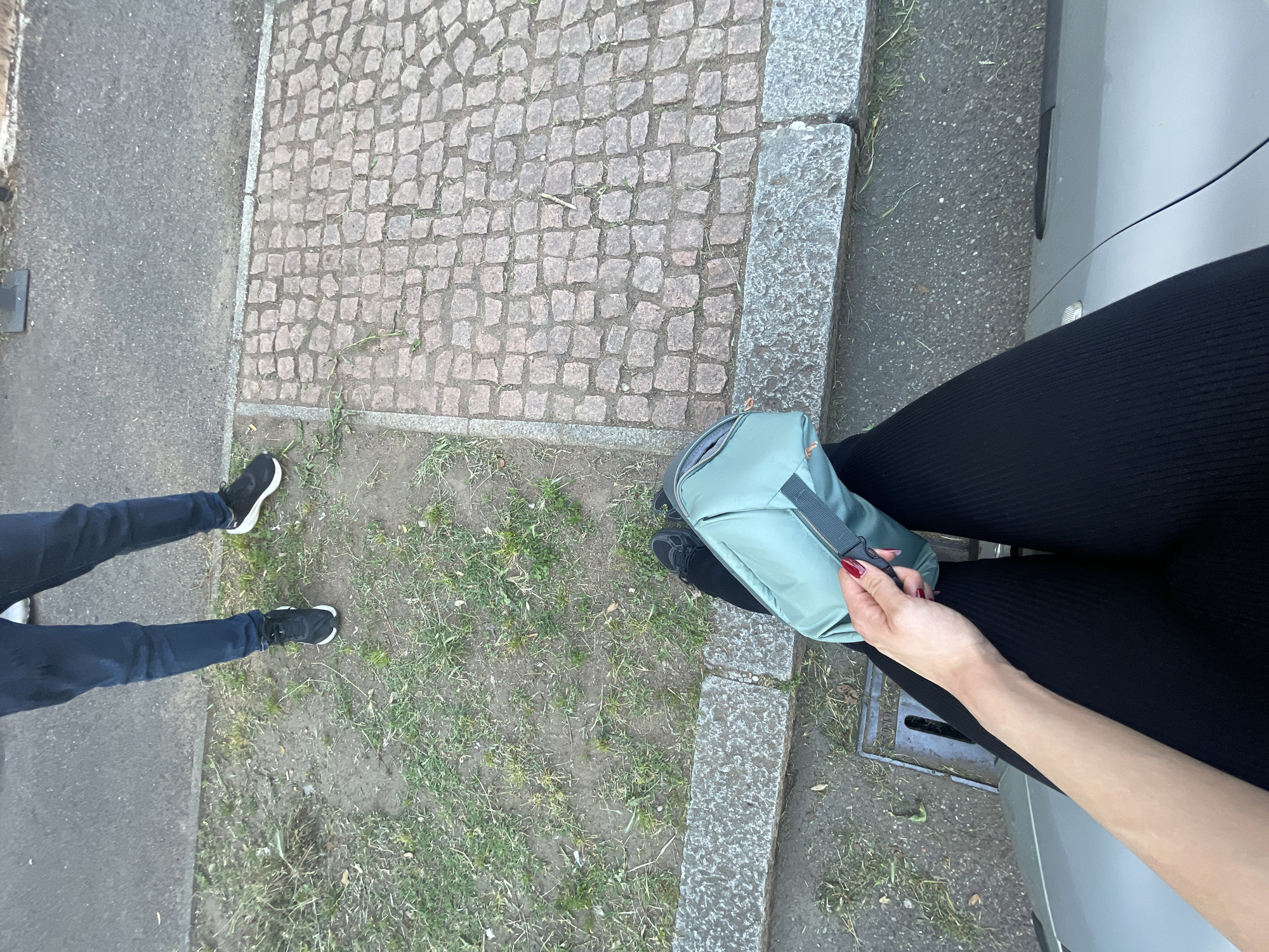 The image depicts a street scene in Busto Arsizio, Italy. The ground consists of cobblestone and asphalt pavement, with patches of grass and weeds growing along the edges. Two individuals are partially visible: one wearing black leggings and the other wearing blue jeans and black shoes. A light blue bag is held by a hand with red nail polish, positioned near the person in black leggings and a parked car. A drain cover is visible near the curb.