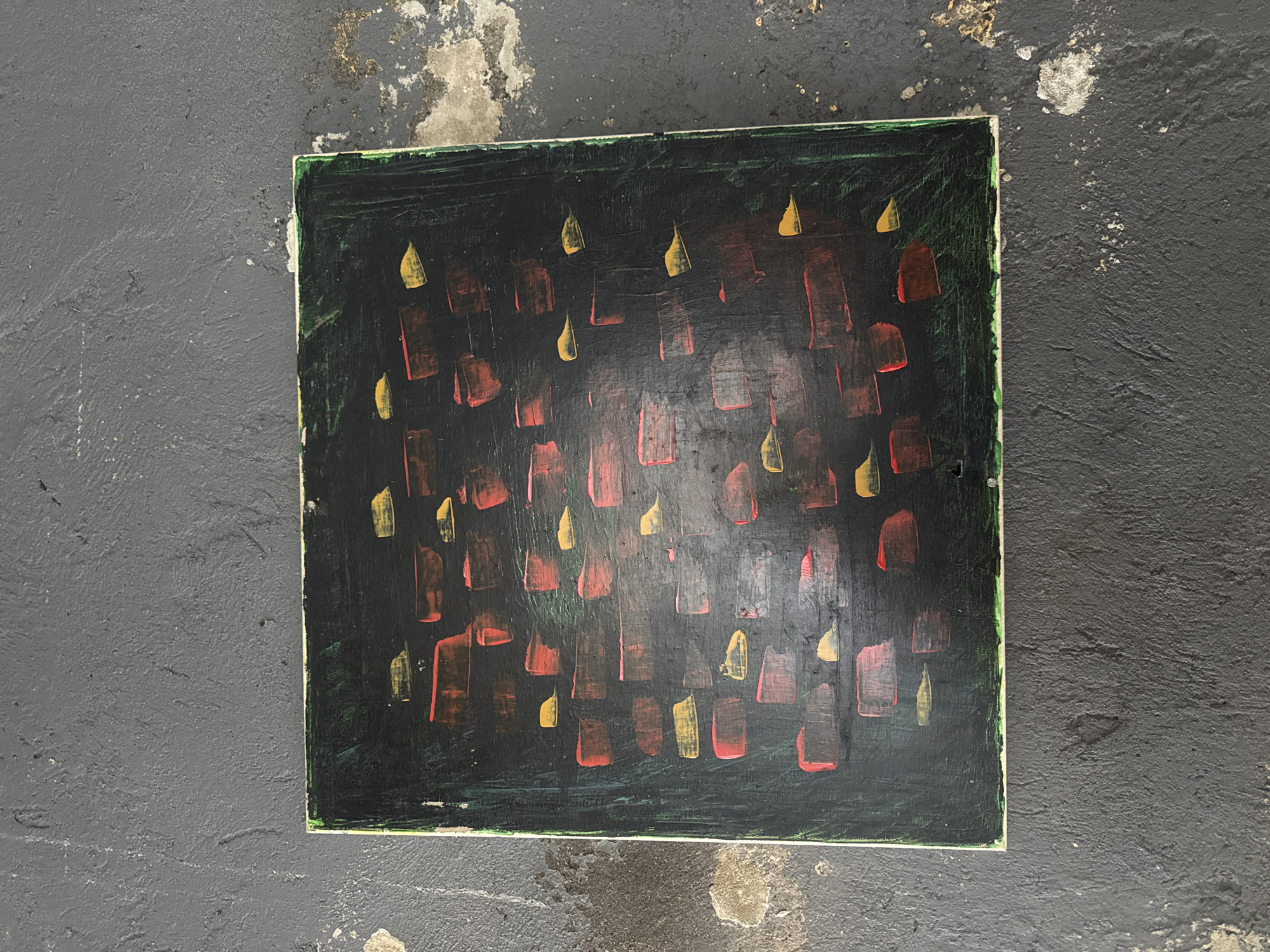 A square, abstract painting with a dark green background is centered on a rough, dark grey concrete surface. The painting features scattered, rectangular brushstrokes in red and yellow, varying in size and density across the canvas. The concrete surface exhibits patches of lighter grey and white discoloration, suggesting wear and weathering. The lighting is consistent with natural daylight, casting soft shadows. There are no people or animals visible in the image. The scene is likely outdoors in an urban environment, given the concrete ground. The specific location of Busungbiu, Indonesia, cannot be determined from the visual information provided in the image.
