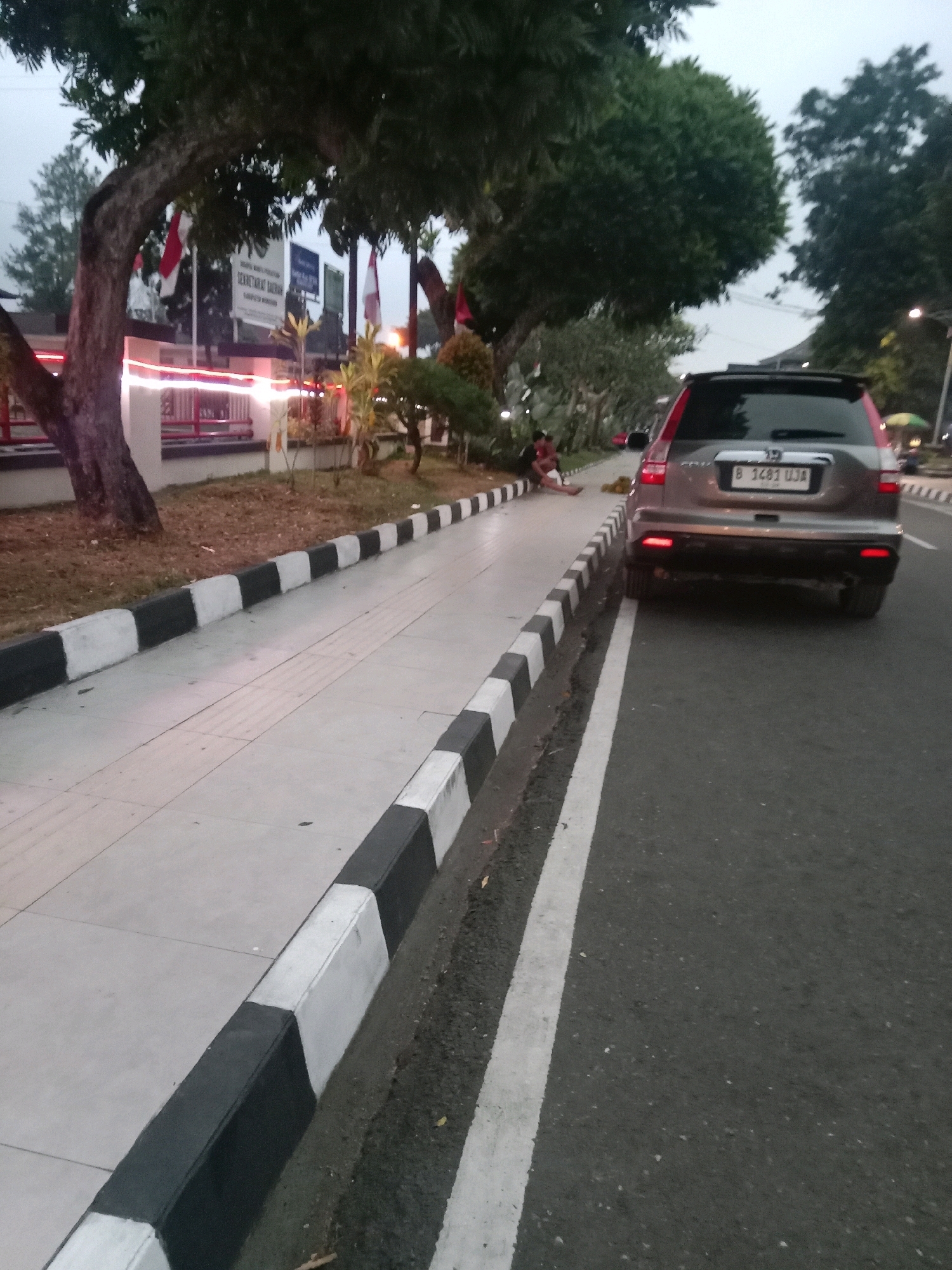 The image depicts a street scene in front of a building in Wonosobo, Indonesia. In the foreground, a paved sidewalk runs alongside a road. The sidewalk has a tactile paving surface and is bordered by a curb painted with alternating black and white stripes. A dark-colored SUV is parked on the right side of the road, facing away from the camera. The SUV has a license plate that reads "B 1481 UJA". On the left side of the sidewalk, a tree with a thick trunk stands near a building entrance with red railings and white decorative lights. Several Indonesian flags are visible in front of the building, along with signage that appears to be in Indonesian. Two individuals are seated on the edge of the sidewalk near the tree, one of whom is holding a child. The environment appears to be an urban or semi-urban setting, with trees lining the street. The sky is overcast, suggesting it may be early morning or late afternoon.