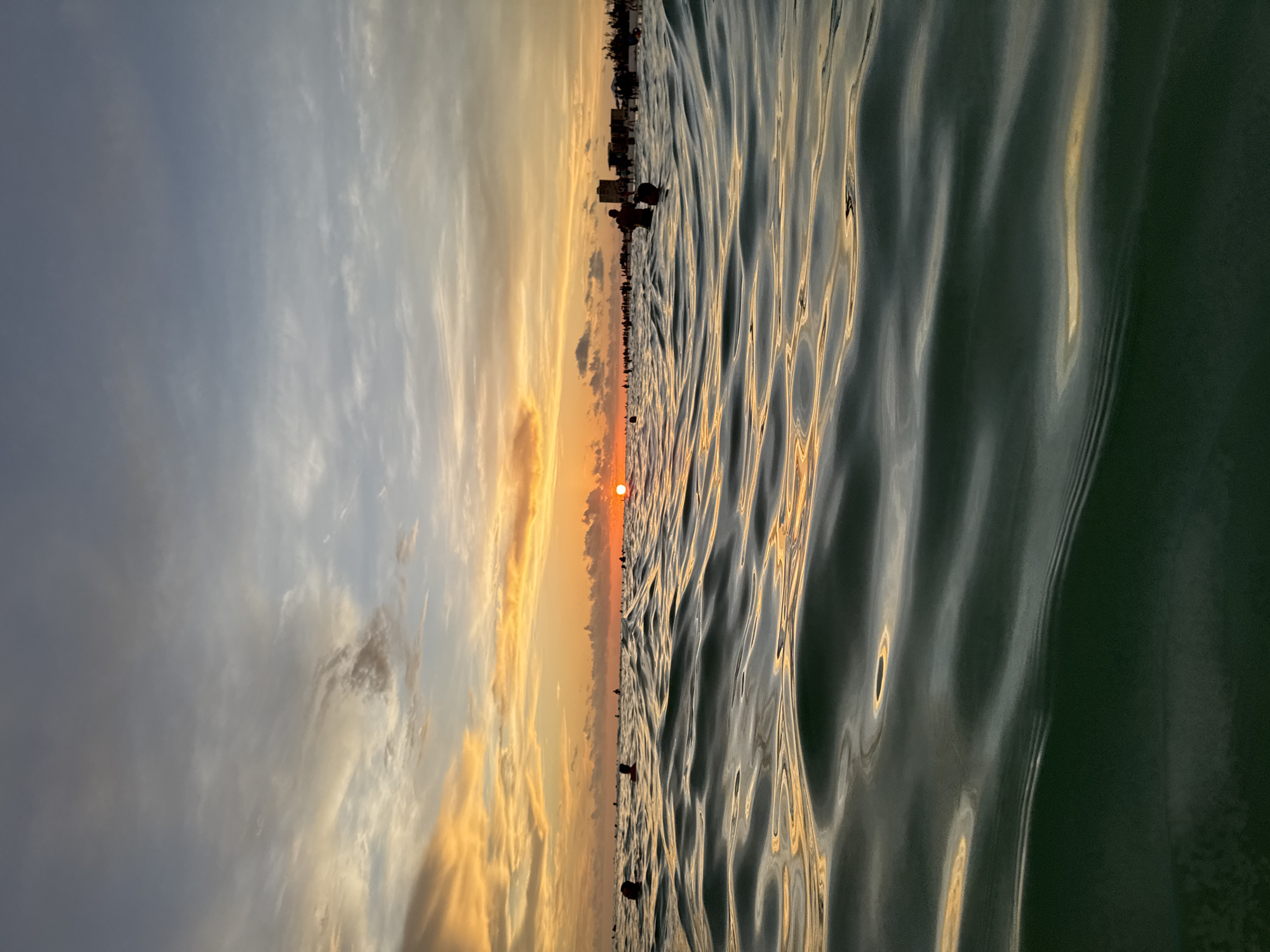 This is a low-angle, wide shot of the ocean at Siesta Key, Florida, during sunset. The foreground is dominated by dark green, choppy water reflecting the warm hues of the sky. In the mid-ground, numerous silhouetted figures are visible in the water, some with their heads above the surface. In the background, a distant shoreline is lined with buildings and a crowd of people, all silhouetted against the vibrant sunset sky. The sun, a bright orange orb, is setting on the horizon, casting a warm glow across the sky, which features streaks of orange, yellow, and blue clouds. The overall scene depicts a lively beach atmosphere at dusk.