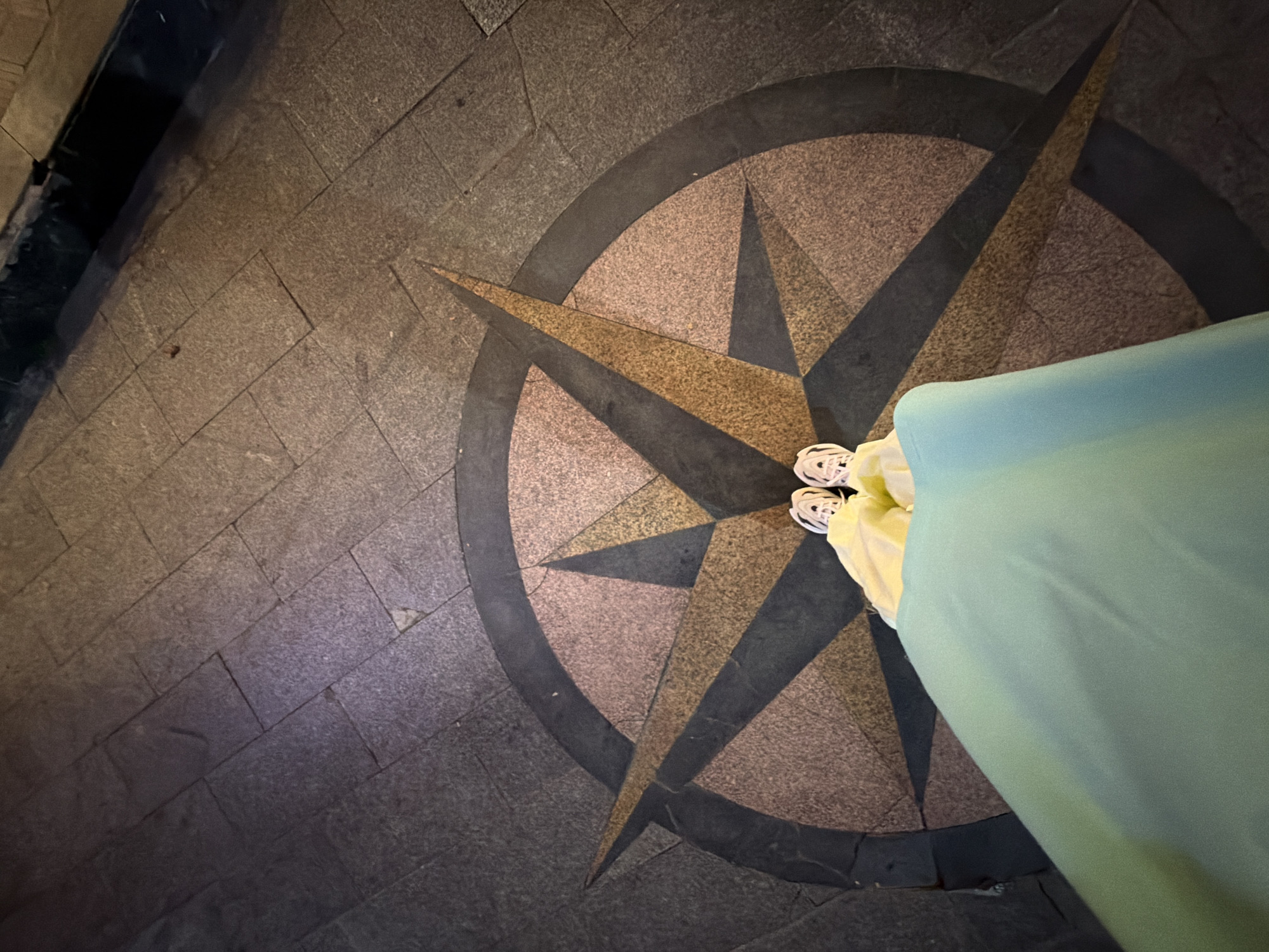 The image shows a close-up, overhead view of a person's feet and lower legs standing on a circular mosaic compass rose embedded in a stone-tiled ground. The person is wearing white sneakers and a light blue garment that covers their legs and extends out of frame to the right. The compass rose has dark grey and tan colored segments, with a prominent dark grey outer ring. The surrounding ground is paved with rectangular, dark grey stone tiles. The lighting suggests it might be evening or nighttime, with artificial lights casting shadows. The scene appears to be an outdoor public space, possibly a pedestrian area or plaza. There is no visible text. The overall impression is one of stillness and perhaps contemplation, with the individual seemingly paused at the center of the compass. The location context of South Quy Nhon Ward, Vietnam, suggests this could be a scenic spot within the city.