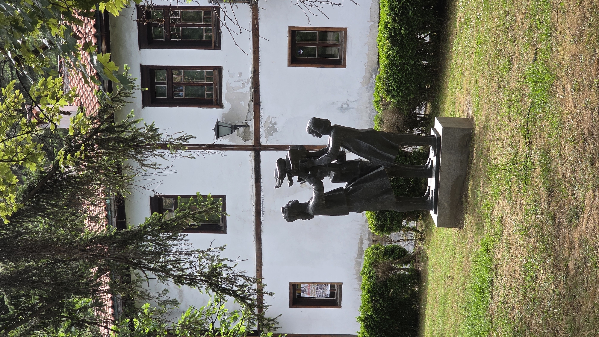 The image shows a bronze statue of a family in front of a white building in Blagoevgrad, Bulgaria. The statue depicts a family of four: two parents and two children. The parents are holding the children up in the air. The statue is set on a stone base in a grassy area. Behind the statue is a white building with brown-framed windows and a red-tiled roof. A small streetlight is mounted to the wall. The building shows signs of wear and tear. The location is likely a park or garden setting in Blagoevgrad.