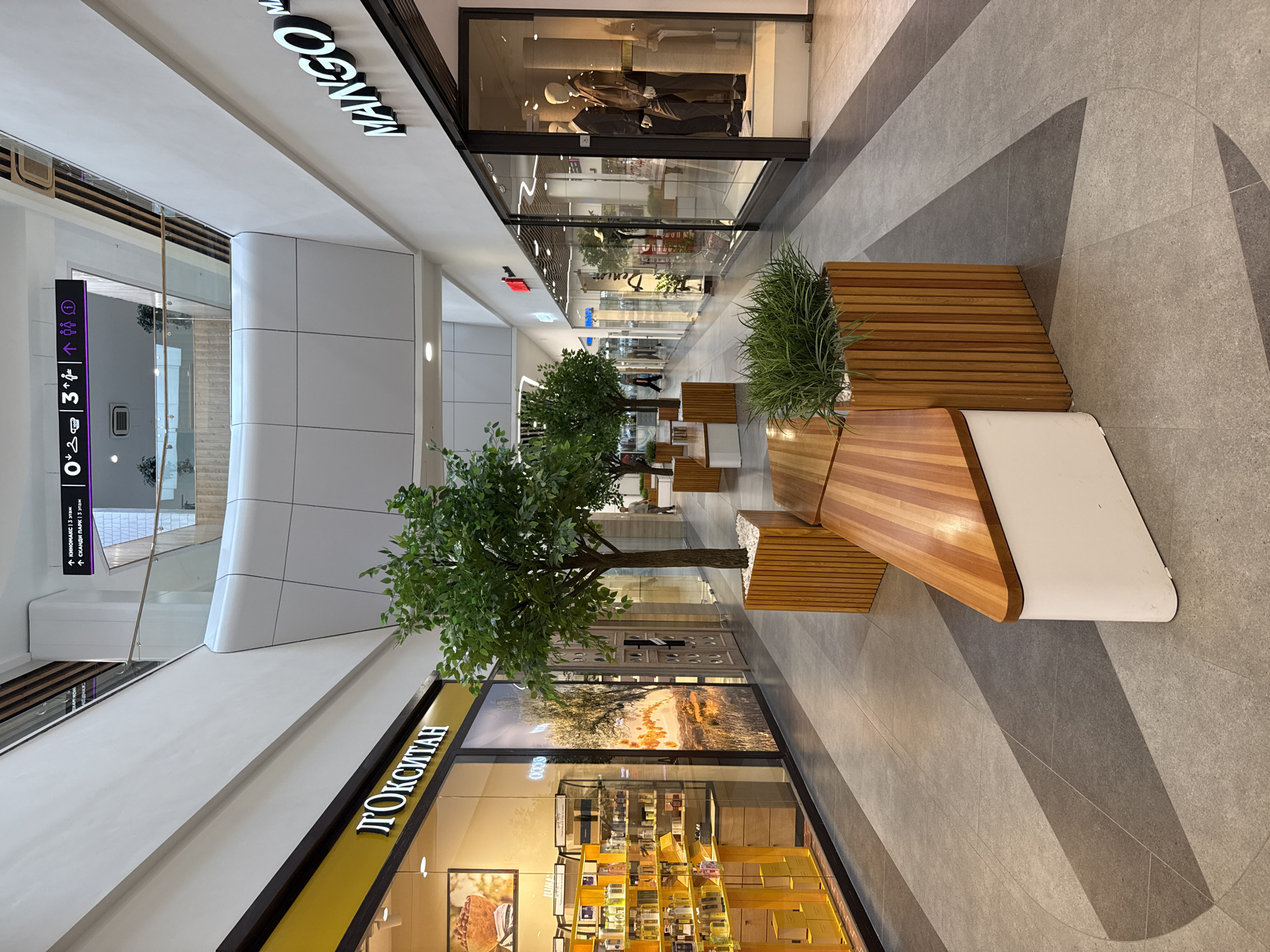 The image shows an interior view of a modern shopping mall, likely located in Izhevsk, Russia, as suggested by the Cyrillic signage. The scene is an indoor walkway with shops lining both sides. On the left, a store with a prominent yellow sign displaying "Л'ОКСИТАН" (L'Occitane) is visible, featuring a display of cosmetic products. Across the walkway, a store with a large "MANGO" logo is visible on the right.
The walkway is paved with large, light gray tiles, with a geometric pattern of darker gray tiles forming curved lines and angles on the floor. In the center of the walkway, there are several modern-looking seating areas composed of wooden benches integrated with large wooden planters containing artificial trees and greenery. These planters are constructed with vertical wooden slats.
In the background, the mall features multiple levels, indicated by a visible upper floor with a glass railing. Overhead, directional signs are mounted on the ceiling. One sign, partly visible, contains Cyrillic text and icons suggesting directions and amenities. The lighting appears to be a mix of overhead fixtures and the illuminated storefronts, creating a bright and clean atmosphere. The overall environment is orderly and well-maintained, typical of a contemporary retail space. No people are prominently featured or actively engaged in any specific interactions. The image captures a static view of the mall's architecture and design.
