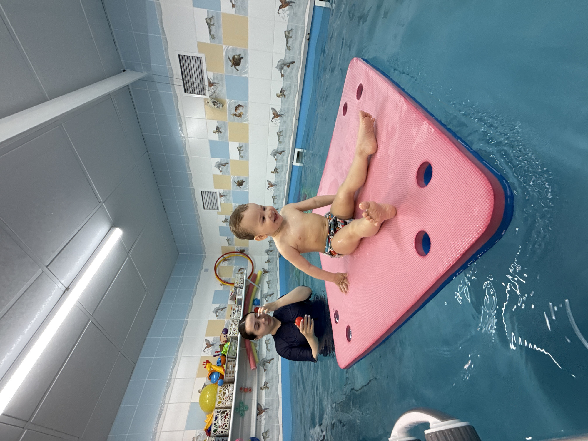 An indoor swimming pool scene captures a happy toddler and an adult engaged in a water activity. A young boy, wearing patterned swim trunks, lies on his stomach on a large, pink floating mat with blue trim, smiling broadly and looking towards the adult. The adult, dressed in a dark long-sleeved swimsuit, is partially submerged in the blue pool water, holding a small red toy, and looking at the child.
The pool area features light blue water and tiled walls. The walls are decorated with a pattern of light blue, white, and beige tiles, adorned with illustrations of aquatic animals such as turtles and birds. Ventilation grates are visible on these walls. Along the wall behind the adult, several shelves hold a variety of colorful swimming toys, including hoops, balls, and plastic shapes. The ceiling is composed of white, square acoustic tiles with bright linear fluorescent lighting fixtures.
The activity suggests a supervised swimming session or playful interaction, possibly an infant swimming lesson, with the child using the floating mat for support. The child's joyous expression indicates a positive and engaging experience in the water. The setting appears to be a child-friendly aquatic facility. No legible text is visible in the image.