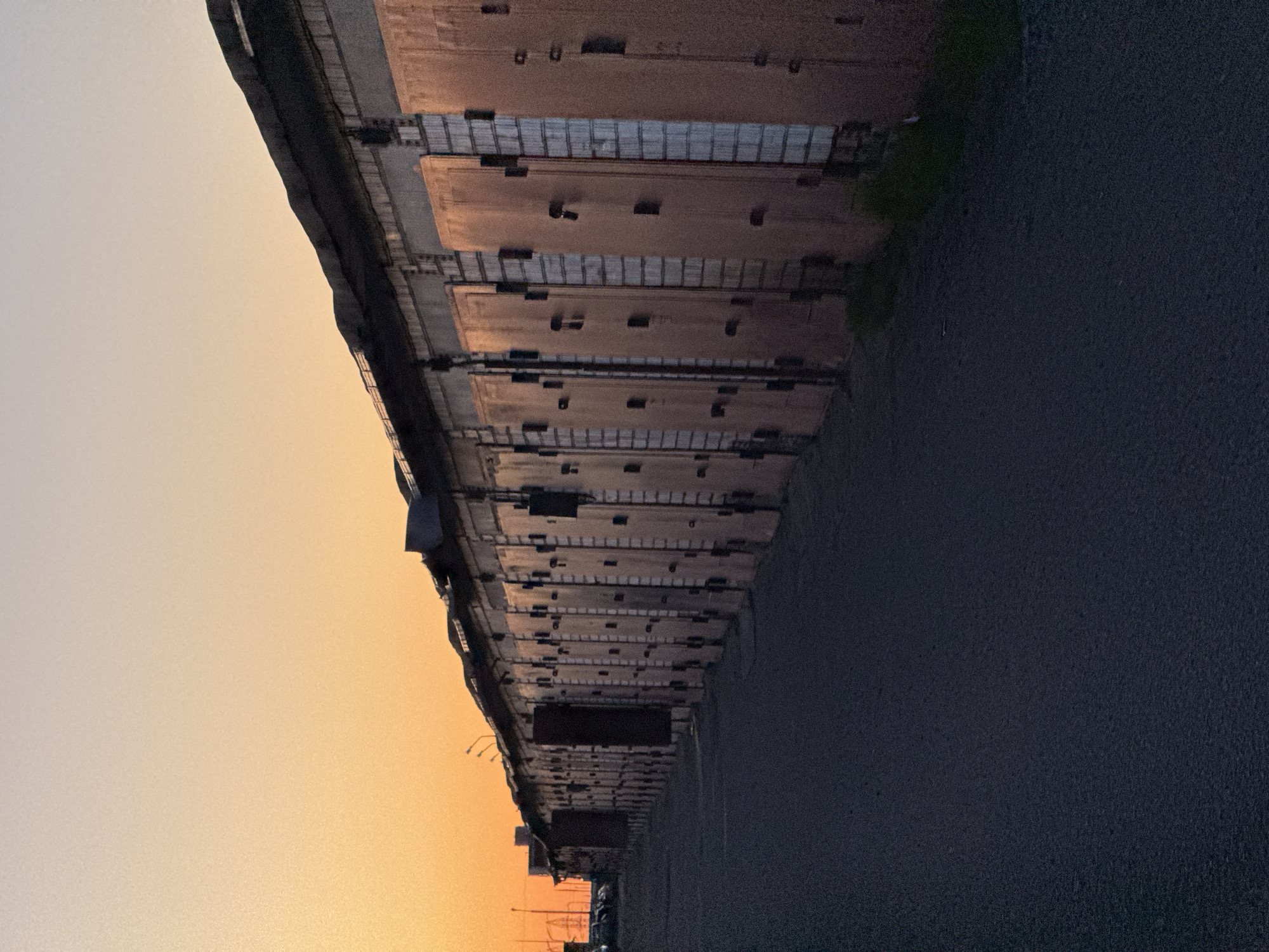 The image shows a long, multi-section building or structure, possibly a parking garage or industrial complex, extending into the distance along a road. The building has a repeating pattern of brown panels with metal support structures between the levels. The sky on the left side is a gradient of orange and yellow, suggesting either sunrise or sunset. The road occupies a significant portion of the lower right area of the image and is dark gray in color. The photograph is taken from a low angle, emphasizing the height and length of the building. The perspective is distorted due to the low angle and possibly the wide-angle lens, causing the building to appear as if it's leaning inwards.