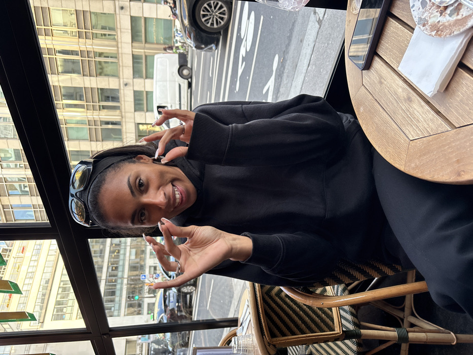 The image captures a smiling young woman sitting at a table inside what appears to be a cafe or restaurant, looking out onto a city street. The picture is rotated 90 degrees counter-clockwise from its natural orientation.
The woman has dark hair, which is pulled back with a black headband, and sunglasses are pushed up onto her head. She is wearing a black hoodie or sweatshirt and black pants. She has a bright, open-mouthed smile, revealing her teeth, and is looking directly at the camera. Her fingernails are long and appear to have a light color, possibly French tips. She is making a similar hand gesture with both hands, where her thumb and index finger are brought together, often indicating "okay" or signifying something small. Rings are visible on her fingers.
To her right, a round wooden table is visible, with a smartphone and a folded white napkin placed on it. Part of a patterned wicker chair is also seen beneath the table.
Beyond the woman and the indoor setting, a large window reveals a bustling city street during the daytime. Tall, modern buildings with numerous windows are prominent across the street, reflecting the light. Cars are visible on the road, including a dark sedan and a white delivery van. White street markings are painted on the asphalt. The overall impression is one of a cheerful moment captured in an urban setting.