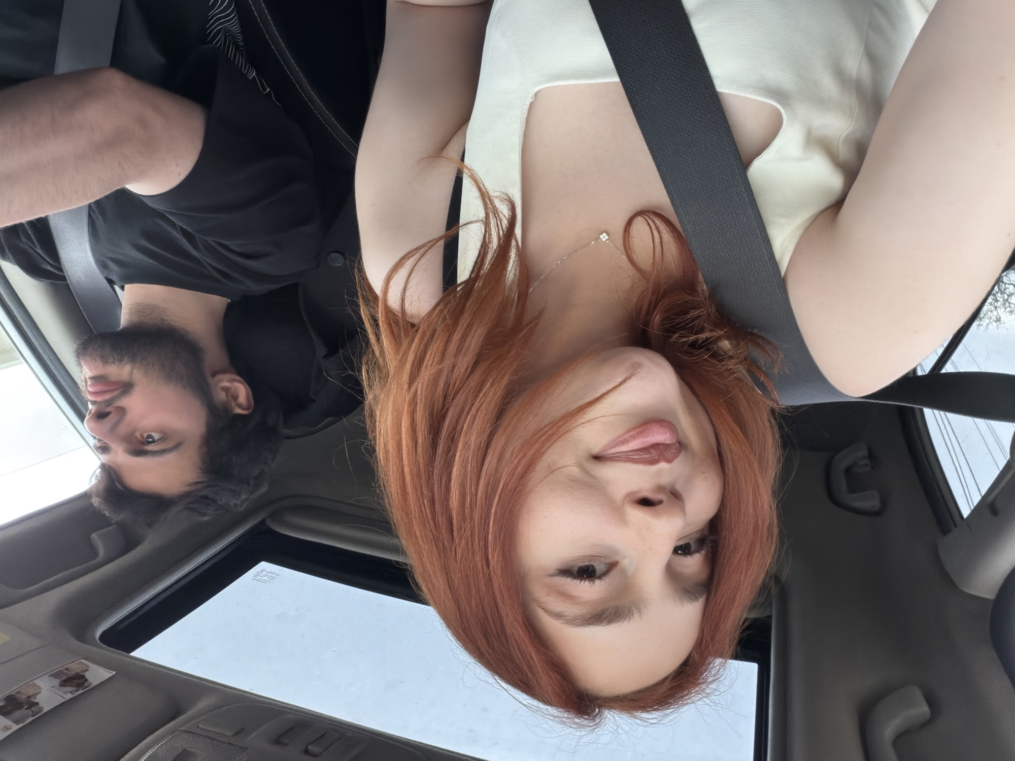 A selfie taken inside a car, likely in Dzegvi, Georgia. Two individuals are visible: a man with dark hair and a beard is partially visible in the lower left corner of the frame. He is wearing a black shirt and a seatbelt. The other person, a woman with reddish-brown hair, is in the foreground, wearing a light-colored top and a seatbelt. She is making a face with her tongue sticking out. The car has a sunroof, visible in the upper left corner. The interior of the car is visible, showing the dashboard and ceiling. The sky is overcast, visible through the sunroof and the window on the right. Power lines are visible through the window.