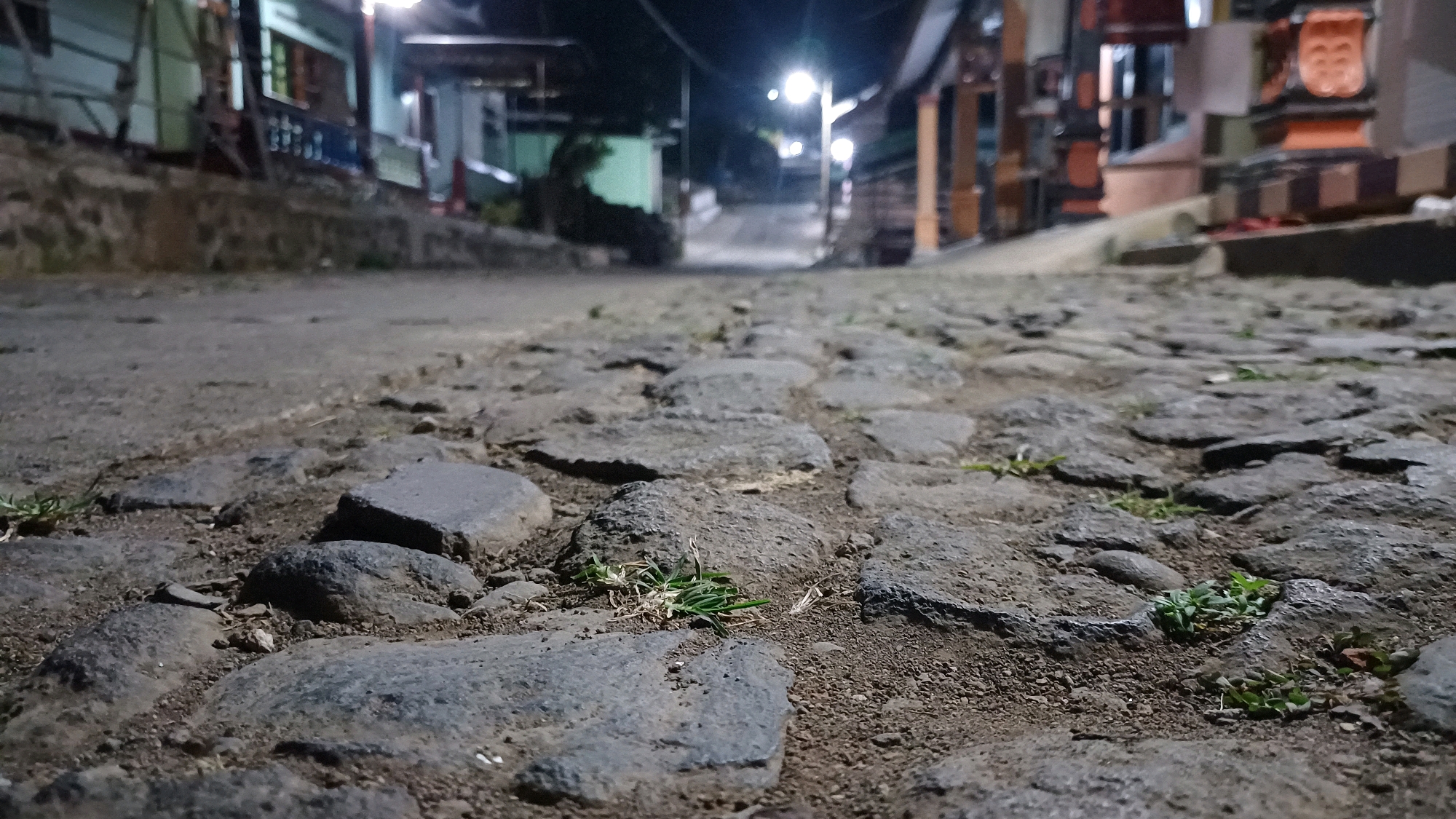The image shows a low-angle, close-up view of a cobblestone street at night in Wonokerso, Indonesia. The foreground is dominated by large, irregular grey stones that form the street surface. Small tufts of green grass sprout between some of the stones. The background is blurred and out of focus, but shows a street lined with buildings. Streetlights are visible, casting a dim glow on the scene. One streetlight in the distance appears particularly bright. The overall impression is of a quiet, dimly lit street in a town or village. There are no people or animals visible in the image. The buildings in the background appear to be a mix of residential and commercial properties, with some architectural details suggesting a traditional Indonesian style.