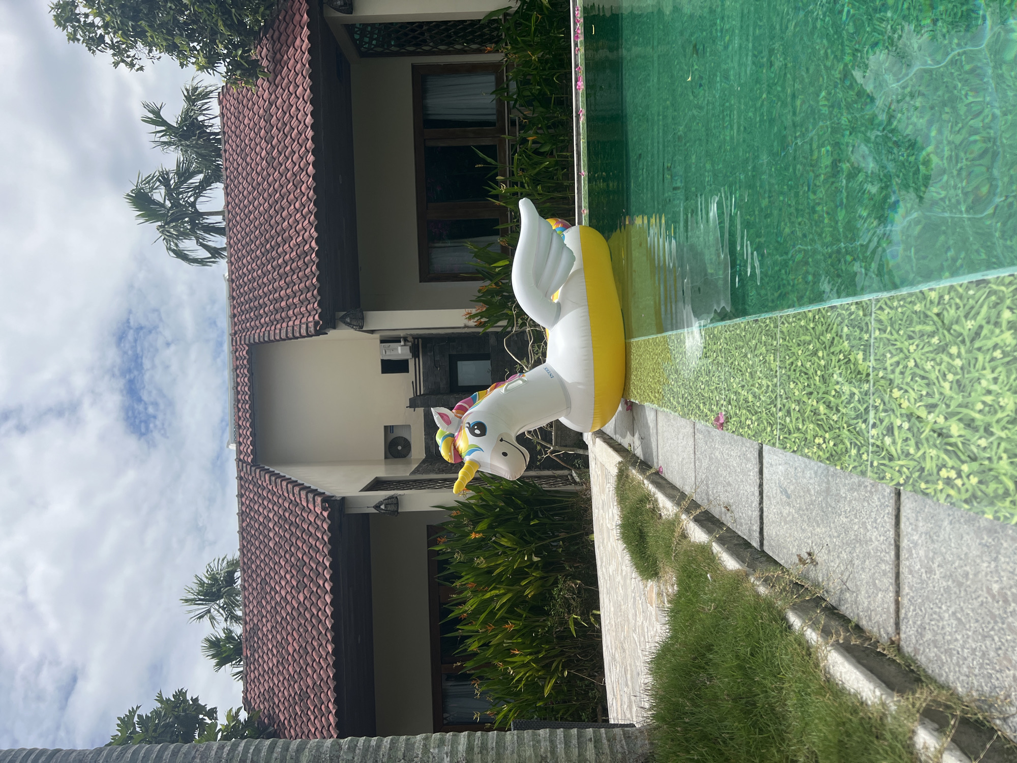 A vibrant, inflatable unicorn float rests partially submerged in a glistening emerald green swimming pool. The unicorn, predominantly white with a bright yellow base, sports a rainbow-colored mane and tail, and a golden horn. Its wings are outstretched, adding to its whimsical appearance. The pool is bordered by a pathway of grey stone tiles and a strip of lush green grass, with a patterned green tile border visible at the edge of the pool.
In the background, a building with a reddish-brown tiled roof and beige walls stands prominently. Large windows with dark wooden frames are visible, partially obscured by dense green foliage, including tall plants with striking orange and yellow flowers. Palm trees with their distinctive fronds sway gently against a sky filled with fluffy white clouds. The overall setting suggests a tropical resort or hotel in Hoi An, Vietnam, given the architectural style and the presence of palm trees. An outdoor air conditioning unit is mounted on the building's exterior. The lighting indicates it is daytime, with the sun casting subtle reflections on the water's surface.
