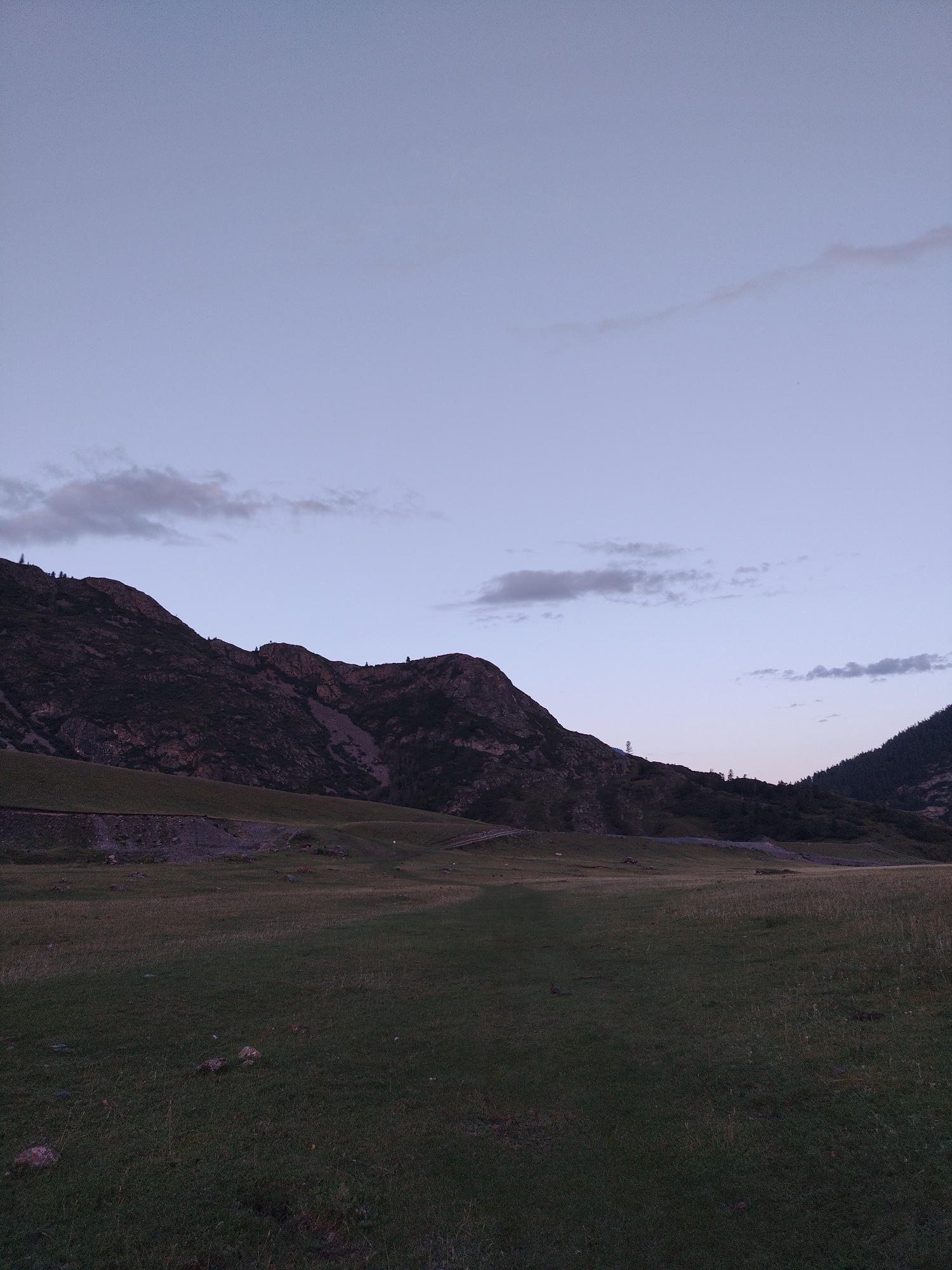 The image depicts a twilight scene in a mountainous, rural landscape, likely near Iodro, Russia. A vast, dark green grassy plain occupies the foreground, sparsely dotted with rocks and tufts of dry grass. In the mid-ground, a gently sloping hill rises, transitioning into a more rugged, darker mountain range that dominates the background. The mountains are characterized by steep, rocky inclines interspersed with darker patches of vegetation and scree slopes. A faint, unpaved road or track is visible winding up one of the lower slopes. The sky above is a pale, hazy blue, with subtle streaks of lighter clouds near the horizon and a few wispy clouds higher up. The overall impression is one of a serene, undeveloped natural environment at the cusp of nightfall. There are no people or explicit human structures, aside from the faint track, visible in the image.