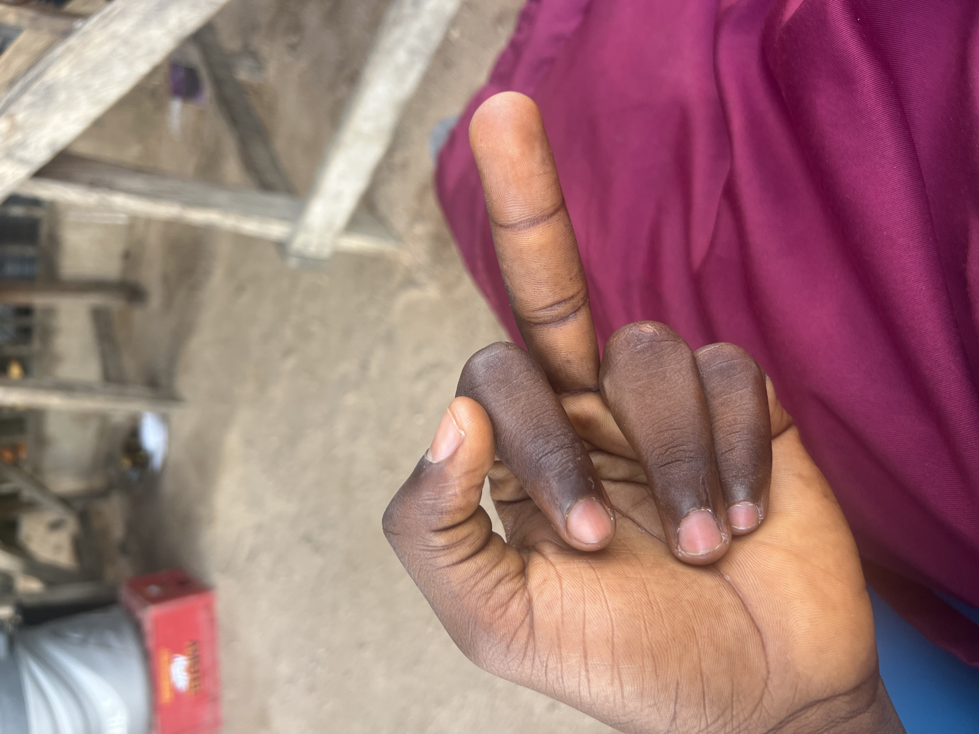 A person's right hand, with dark skin, is prominently displayed. The middle finger is extended upwards in an offensive gesture. The person is wearing a maroon shirt or garment. The background is slightly out of focus, showing what appears to be the edge of a building, an outdoor setting with a sandy ground, and a partially visible red object. The location of the image is in Jalingo, Nigeria.
