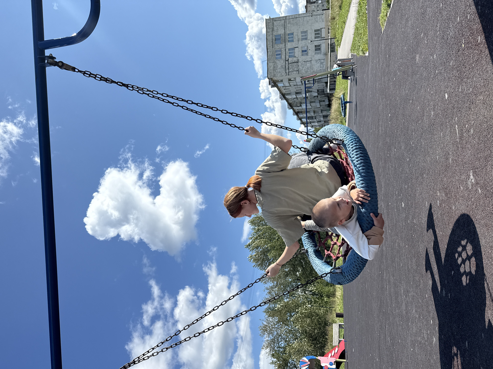 In the city of Gornozavodsk, Russia, on a sunny day, two children are enjoying a swing set. A young girl with auburn hair pulled into a ponytail is seated on a large, circular blue swing, holding onto the chains. Beside her, a young boy in a white long-sleeved shirt is lying down on the swing. The swing set is situated on a black rubberized playground surface. In the background, a line of green trees can be seen, followed by a weathered, multi-story concrete apartment building. The sky is a vibrant blue, dotted with white, puffy clouds. The overall scene depicts a typical outdoor recreational area in a residential district.