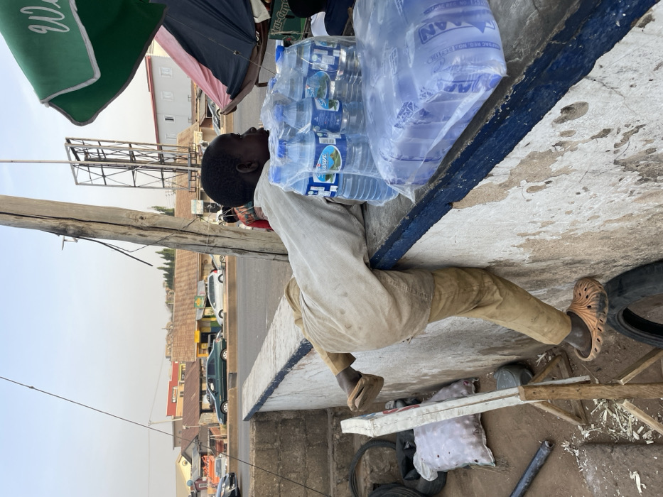 A man with dark skin carries a heavy load of bottled water on his head, balancing it precariously. He wears a light-colored, long-sleeved shirt and tan pants, with sandals on his feet. He is perched on a concrete ledge near a busy street with cars and buildings visible in the background. A green canopy and other objects clutter the foreground. The image appears to be taken outdoors on a sunny day.