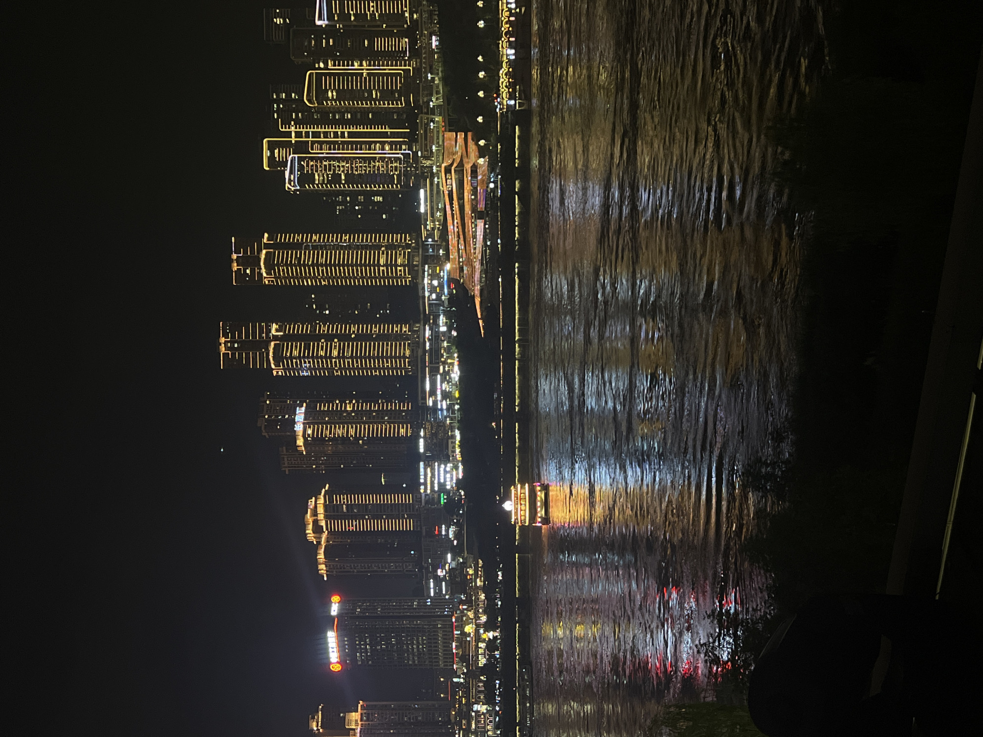 The image captures a nighttime cityscape along a riverfront, likely in Jiangbeicheng Street, China. A large river dominates the foreground, its surface reflecting the illuminated buildings and lights from the city. The water has a textured, rippling appearance, with streaks of light creating a dynamic visual effect.
In the middle ground, a multi-decked passenger boat with numerous illuminated windows is visible on the river, appearing to be in motion. Along the riverbank, a collection of modern buildings and skyscrapers are brightly lit, their facades adorned with intricate patterns of yellow and white lights. Some buildings have distinctive architectural features, with layered structures and illuminated outlines. One prominent building exhibits a unique, undulating design with colorful light projections.
The background consists of the dark night sky, with the city lights creating a glowing horizon. The overall impression is a vibrant and dynamic urban scene at night, showcasing the architectural splendor and the lively atmosphere of the riverfront. No people are clearly discernible in the image.