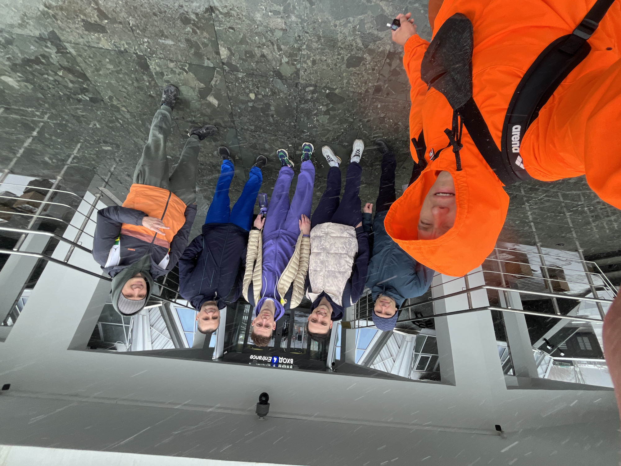 Five individuals stand near a reflecting surface, likely a polished marble floor, creating a distorted, inverted reflection of the building and surroundings. The reflection indicates the location is near the entrance of the Zaryadye Park Media Center in Moscow, Russia. The individuals are dressed in cold-weather attire, suggesting a cool or cold temperature. One individual wears a bright orange hooded jacket with a black shoulder bag. Others wear various jackets and pants in shades of blue, purple, and gray. The reflection also captures elements of the building's architecture, including windows, railings, and signage indicating the entrance. It appears to be snowing.