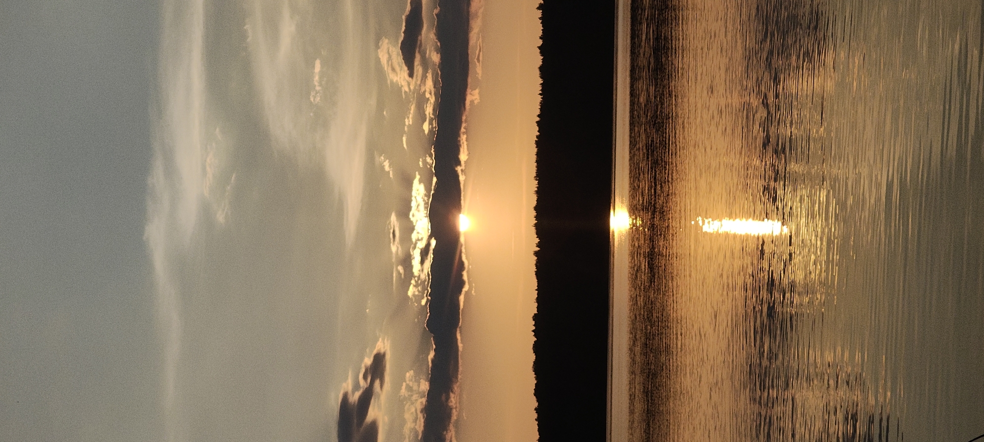 The image depicts a sunset over a body of water, likely a lake, in the vicinity of Mednoye Ozero, Russia. The sky is filled with various clouds, some dark and ominous, others wispy and illuminated by the setting sun. The sun itself is partially obscured by a thick band of dark clouds, casting a warm, orange glow across the sky. A reflection of the sun creates a shimmering path on the rippling surface of the water. A silhouetted treeline forms the horizon, separating the sky from the water. The overall scene is one of natural beauty and tranquility.