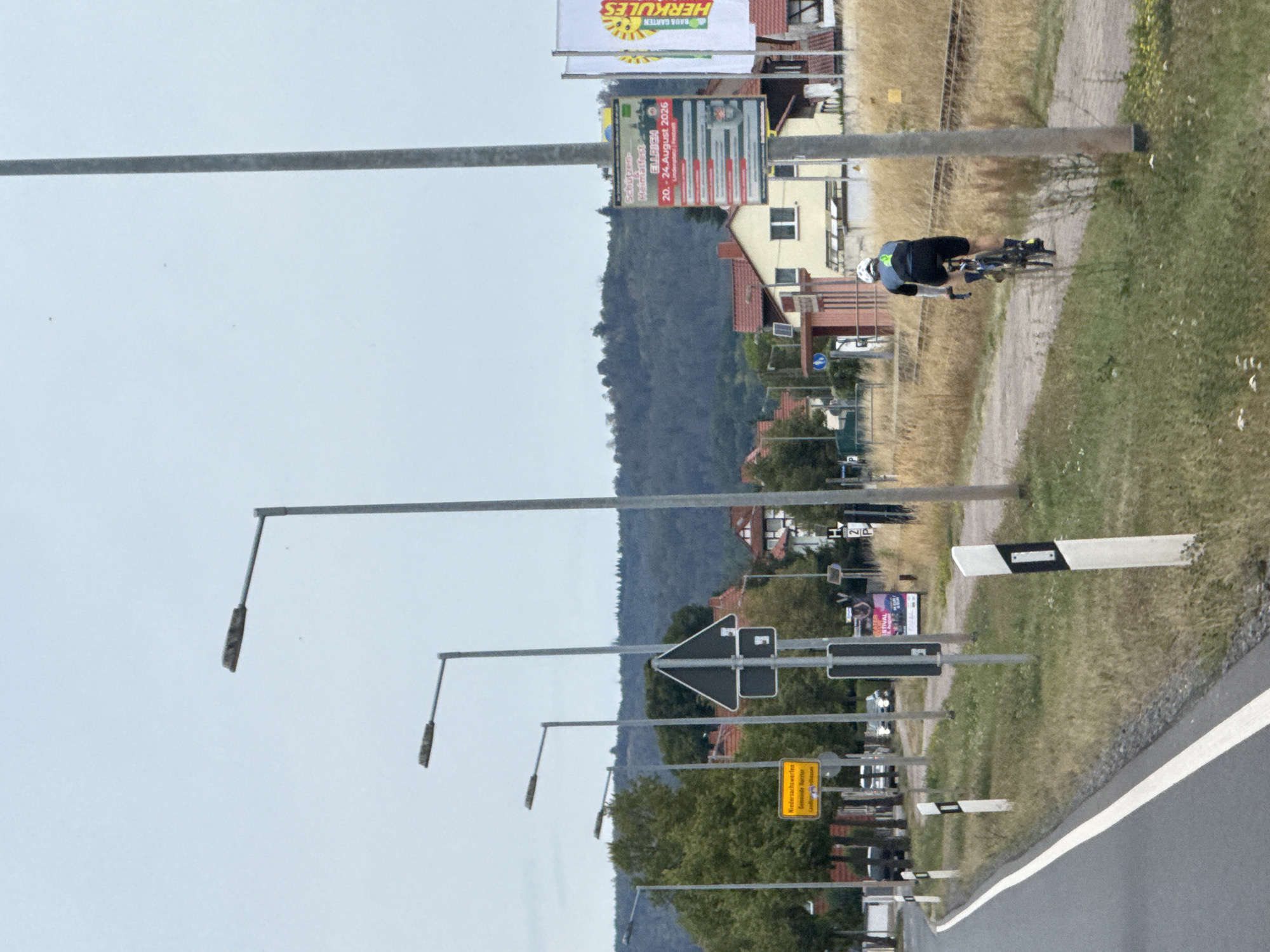The image depicts a street scene in Niedersachswerfen, Germany. A cyclist wearing a white helmet, dark blue and gray cycling attire, and black shorts is riding away from the camera on a gravel path. The cyclist is positioned to the right of the frame, with a tall, gray lamppost to their right. In the background, there are houses with red roofs, trees, and a forested hill under a cloudy sky. Several lampposts are visible along the street, and a yellow sign with German text is mounted on a pole on the left side of the road. A white and black striped marker post is located in the foreground on the right. A larger sign with event details, including dates "20.-24. August 2026," and two flags, one yellow and one white with text that includes "HERKULES," are also visible near the lamppost and cyclist. Train tracks run horizontally in the mid-ground behind the gravel path.