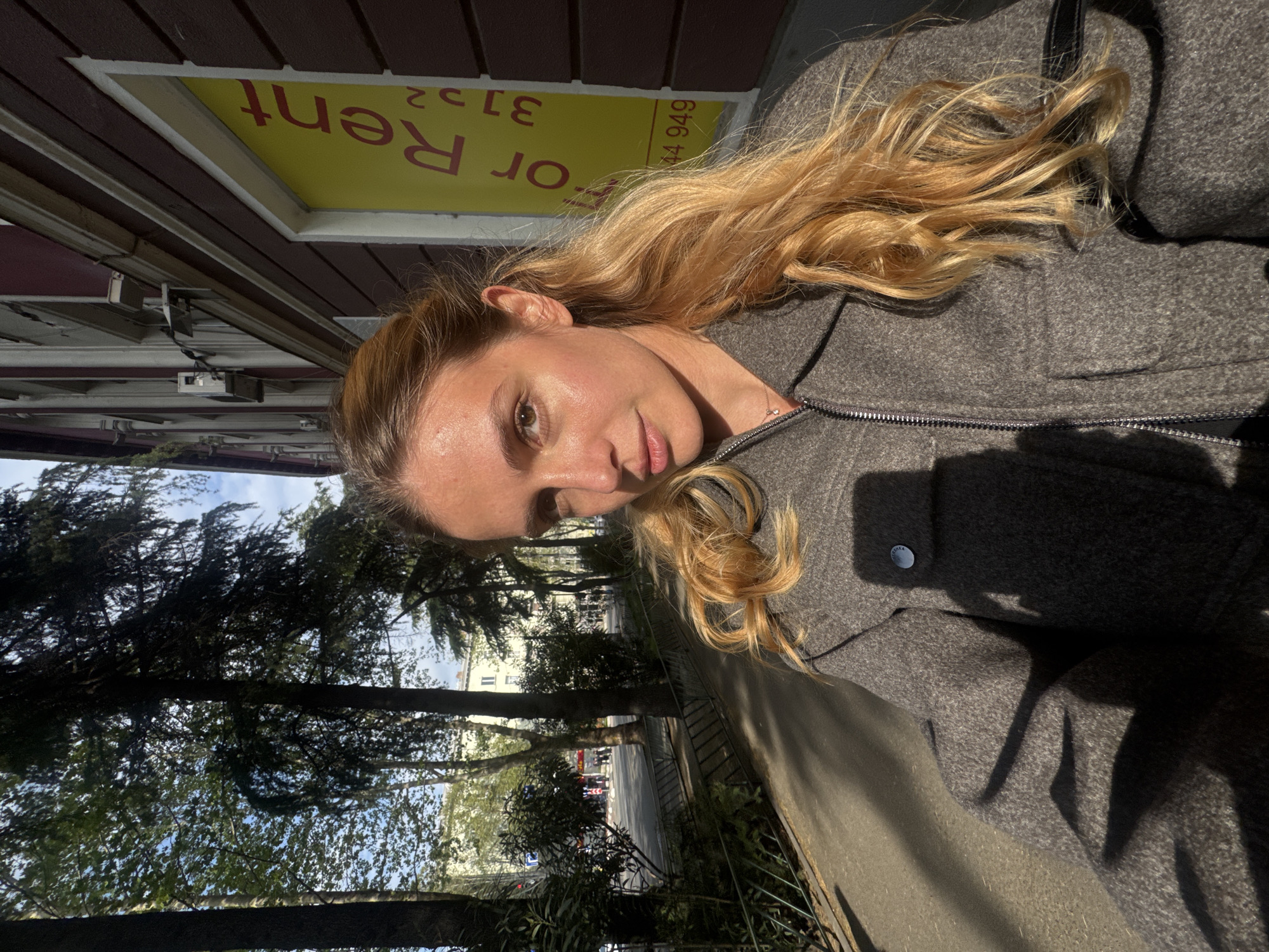 The image features a young woman outdoors in a sunlit setting. She has light skin, brown eyes, and long, wavy blonde hair, looking directly at the camera with a calm, slight smile. She is wearing a grey zippered jacket or coat, with a small silver button visible on one sleeve.
Behind the woman, a building with reddish-brown horizontal siding is visible. In one of its windows, a yellow "For Rent" sign is prominently displayed, showing the text "For Rent" (partially reversed, suggesting it's seen through glass) and partial phone numbers "3132" and "44 949". To the woman's left, a row of tall trees with dense green foliage frames the background, and beyond them, a street with distant buildings and possibly a vehicle can be seen under a partly blue sky.
The bright sunlight indicates a clear, sunny day, casting distinct shadows and highlighting parts of her hair and face. The overall scene suggests an urban or suburban street in Tbilisi, Georgia, combining architectural elements with natural greenery. The woman is the main subject, appearing relaxed in the daytime environment.