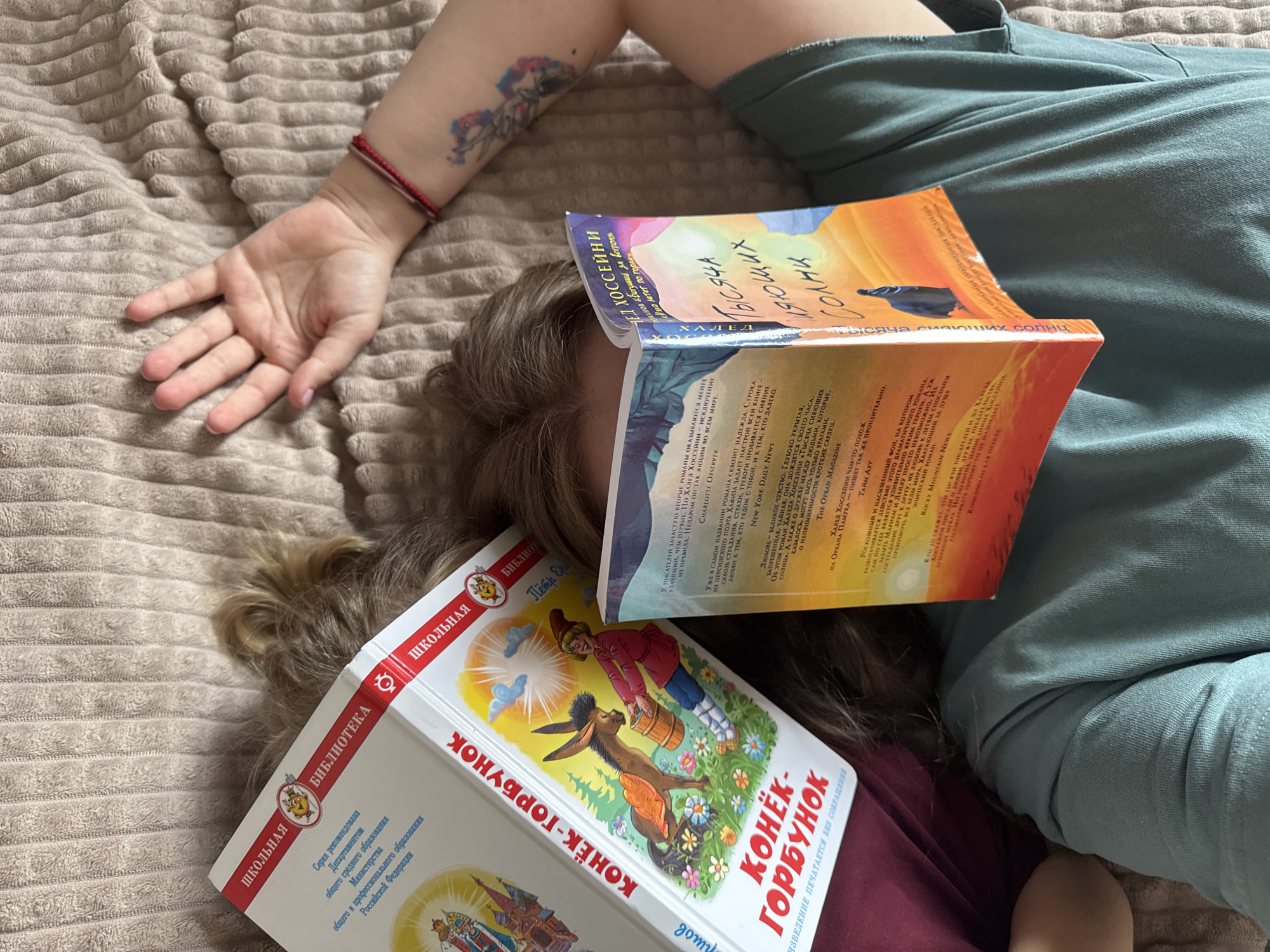 A person is lying on a tan textured surface, possibly a bed or couch, with two books covering their face. The books are a children's book titled "Konek-Gorbunok" and a novel titled "A Thousand Splendid Suns". The person's right arm is extended with their hand open, and a tattoo is visible on their forearm. They are wearing a red bracelet. The person is wearing a green shirt and a maroon shirt underneath. Their hair is light brown and wavy. The setting seems to be indoors. The location is not Gornyi (Dvurechenskoe S/P), Russia because that is not visible in the image.