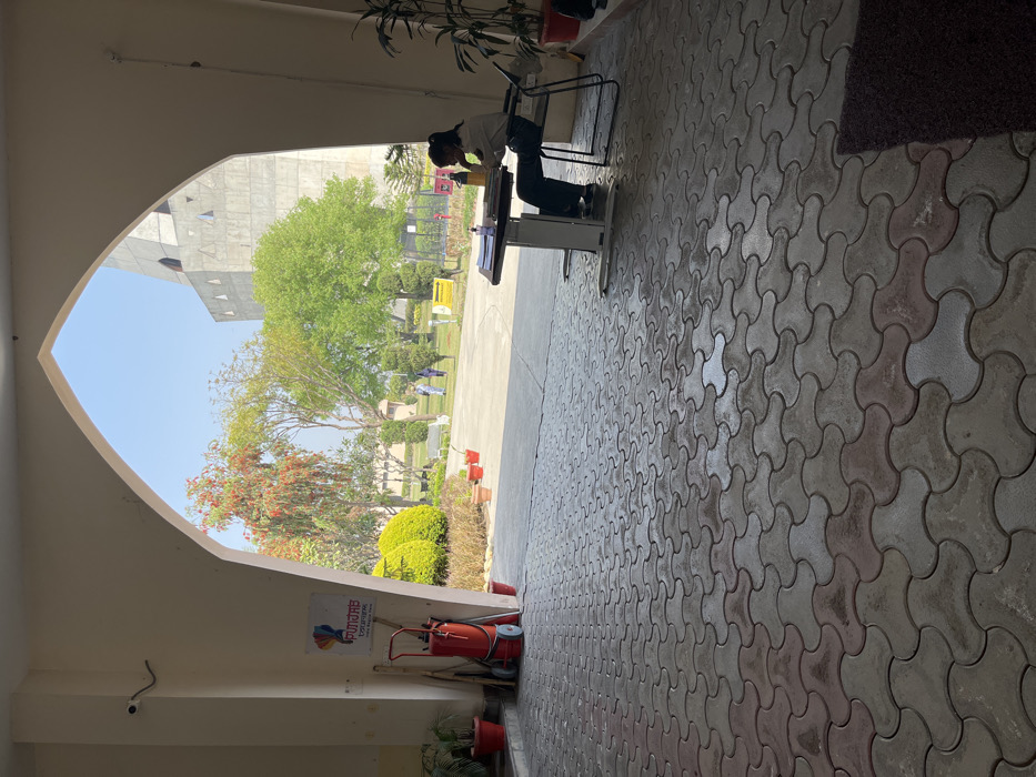 The image shows a view from inside a building in Sahibzada Ajit Singh Nagar, India. The view is through an arched doorway, revealing a paved courtyard with a person sitting at a desk. The person appears to be writing in a notebook and holding a water bottle. There are several potted plants surrounding the area. There is a sign on the left wall that reads "Punjab Tourism," in a multicolor font, and a fire extinguisher attached to a cart. Beyond the courtyard, there is a large, grey concrete structure visible in the distance, partially obscured by lush green trees. The sky is a bright blue with no clouds. The paving is a dark grey, with a pattern of irregular interlocking pieces. There are several other people, obscured in the distance, walking on the paved path. The image was captured during the daytime, in what appears to be pleasant weather.