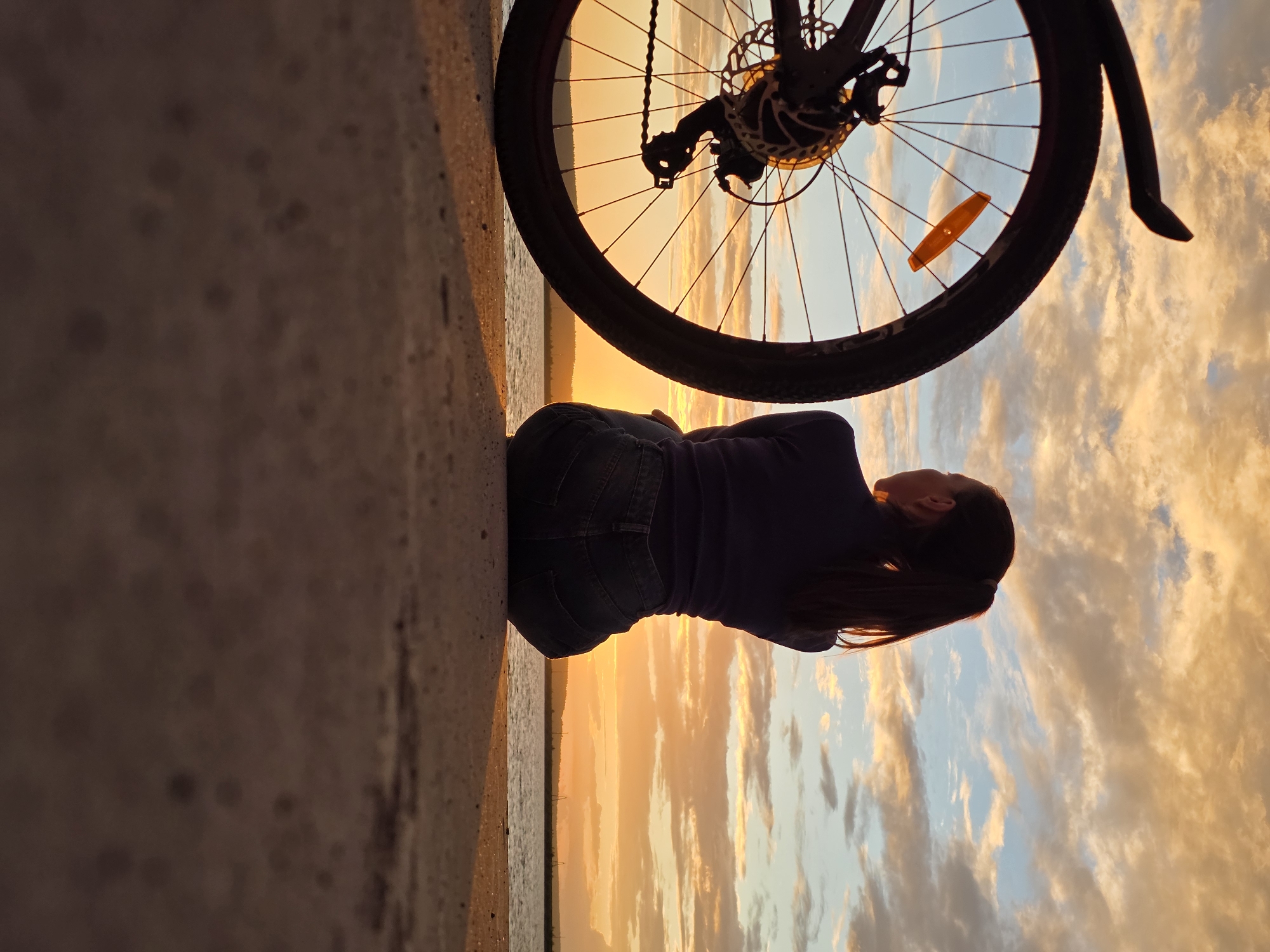 The image shows a person lying on a concrete surface with a bicycle wheel obscuring part of the view. The person is wearing jeans and a dark top and is facing away from the camera, looking towards a sunset over a body of water. The sky is filled with dramatic, colorful clouds, indicating either sunrise or sunset. The concrete surface appears to be an embankment or pathway. The scene suggests a moment of rest or contemplation during an outdoor activity, possibly cycling. The time of day is clearly sunset due to the warm, golden hues of the sky and clouds. The location cues are not distinct enough to pinpoint a specific area within Chaikovsky, Russia, but the presence of water and a pathway is evident. There is no visible text in the image. The overall mood is peaceful and serene.