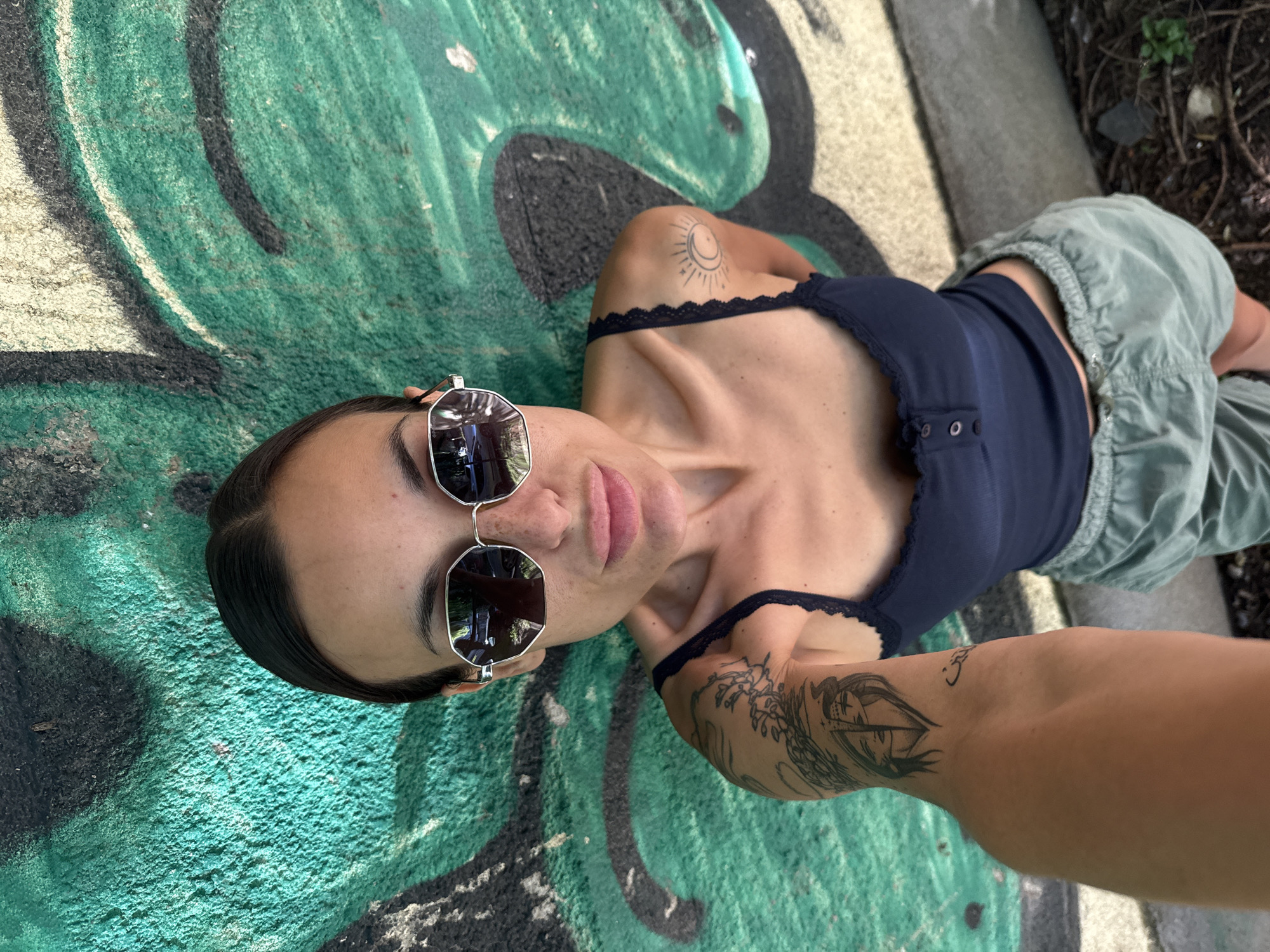 A young woman is captured in a close-up, low-angle shot, lying on her back on a vibrant, graffiti-covered concrete surface. She looks directly at the camera with a neutral expression, shielded by reflective, hexagonal-framed sunglasses. Her dark hair is neatly pulled back, and she wears a dark blue camisole with delicate lace trim along the neckline. Her attire includes light green or grey cargo pants, visible on her lower body.
Several tattoos adorn her arms: a sun and crescent moon design is visible on her left shoulder, and a larger, intricate, stylized tattoo covers her right forearm, depicting what appears to be an eye or a boat. Another small, simple tattoo is on her right wrist. The backdrop is characterized by bold, abstract graffiti in shades of bright green and black, creating a textured and urban aesthetic. To the right of the frame, glimpses of natural ground cover and sparse foliage suggest an outdoor environment. The bright, even lighting indicates it is daytime, likely sunny. The woman appears relaxed and at ease in this outdoor setting, which is located in Pleven, Bulgaria. No visible text is present in the image.
