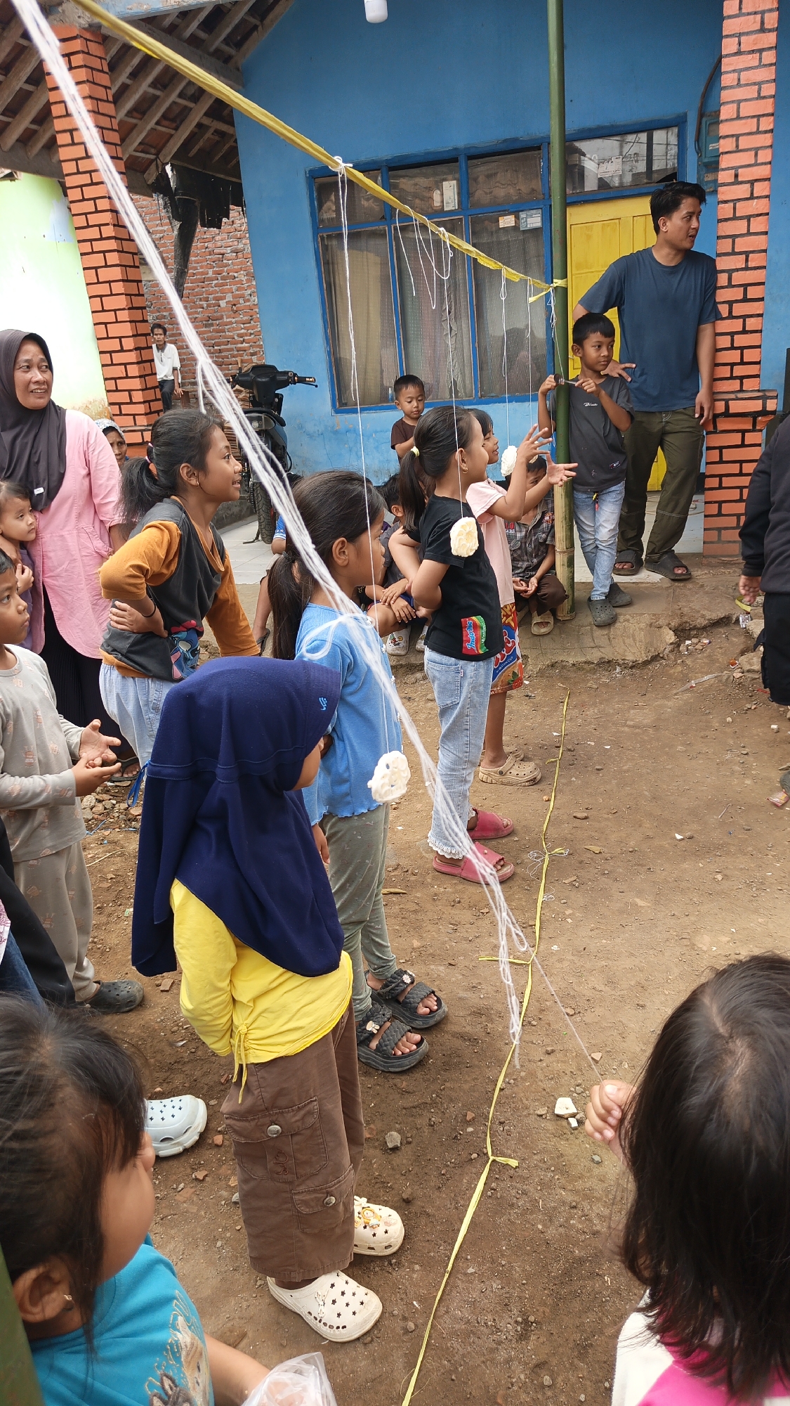 In Linggasirna, Indonesia, a group of children are participating in a game involving food items suspended on strings. A blue building with orange brick accents serves as the backdrop. The ground is dirt and uneven.
Several children, mostly girls, are actively engaged in the activity. They are dressed in casual attire. One girl, wearing a blue hijab and a yellow shirt, stands in the foreground. To her right, another girl in a blue shirt reaches out towards the suspended food. A girl in a black shirt with a colorful graphic on it is also reaching for the food. To the left, a girl in a gray shirt and light-colored pants is looking towards the game.
Adults are also present. A man in a dark blue t-shirt and green pants stands on the right, observing the children. A woman in a pink top and black hijab is on the far left, also watching.
The game involves strings, some of which have white, puffy food items attached. A yellow ribbon is laid out on the ground, possibly marking a boundary or course. A motorcycle is parked in the background. The overall scene depicts a community gathering or celebration, likely a children's event.