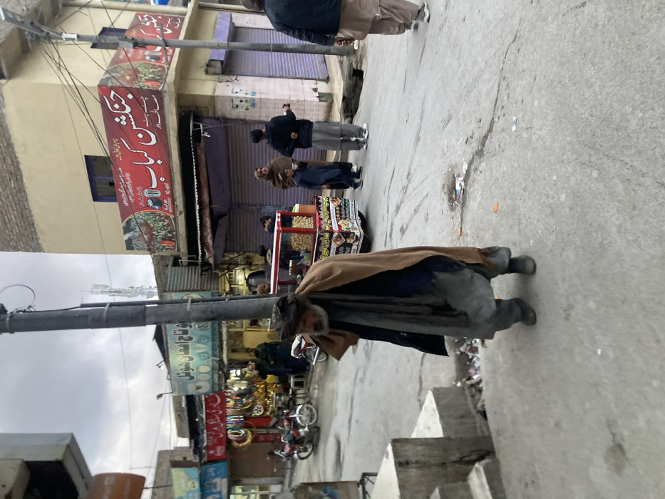 An elderly man in a worn coat walks along a gray street. Shops line the street, with signs in a script, possibly Urdu. Several people stand by a food cart, while a few others stroll by. The sky is overcast. A utility pole stands to the left of the man.