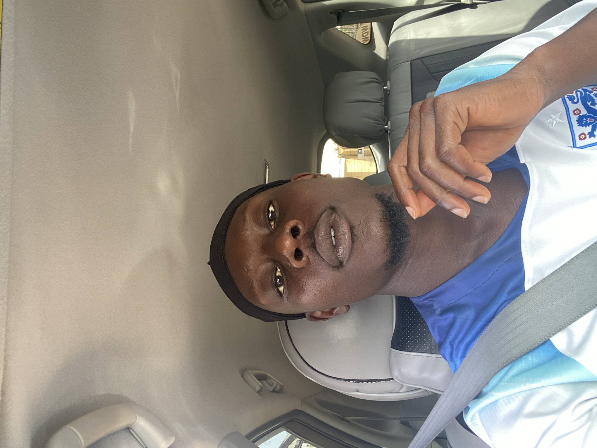 A dark-skinned male wearing a black skullcap and a white and blue soccer jersey is pictured inside a vehicle. He is looking directly at the camera with his hand resting on his chin. The interior of the vehicle is visible, including seats and the headliner. The male is seated in the driver's seat, indicated by the seatbelt diagonally across his chest. The background outside the car is blurred, but shows buildings and vegetation. The image appears to be taken during daylight. The location is stated to be Bar Jirgi Summa, Nigeria.