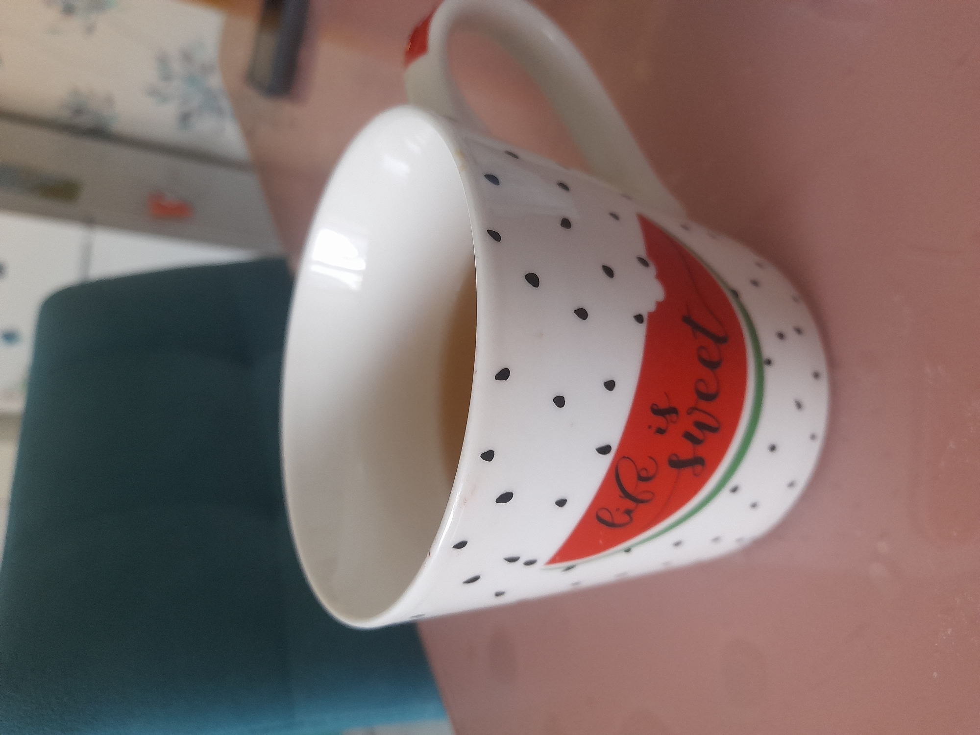 The image shows a white ceramic mug with black polka dots and a red watermelon design with the text "Life is sweet" in black cursive. The mug is sitting on a pink surface. A green chair is visible in the background.