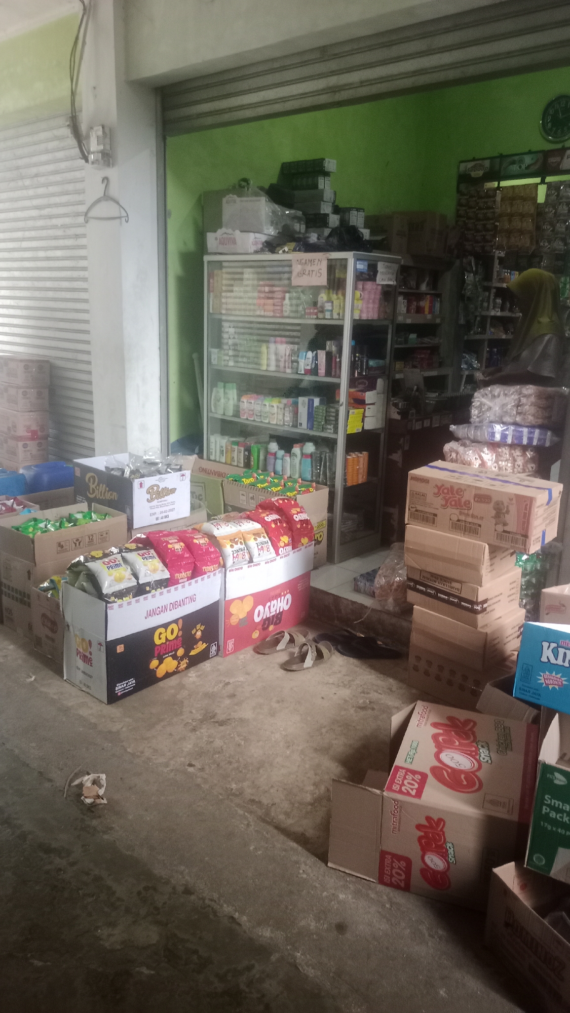 The image depicts a retail store, likely a small grocery or convenience store, located in Cogreg, Indonesia. The store appears to be cluttered with various products, primarily food items and household goods. In the foreground, several cardboard boxes containing snacks are prominently displayed. One box, labeled "GO! PRIME," features images of chips. Another box, larger and black and white, also advertises "Billion" snacks. To the right, a stack of boxes labeled "Yale Yale" and "Copla" are visible.
Behind the snack displays, shelves filled with bottles and packaged goods can be seen, suggesting a variety of products are for sale. The store's walls are painted a vibrant green in the back. The flooring is a rough, concrete surface. A closed metal shutter is visible on the left side of the image, indicating the store's entrance or a part of its facade. A clothes hanger is attached to a vertical pillar near the shutter. In the background, a person wearing a hijab is partially visible, suggesting a shopkeeper or customer. The overall impression is of a modestly stocked, functional retail space in a local Indonesian community.