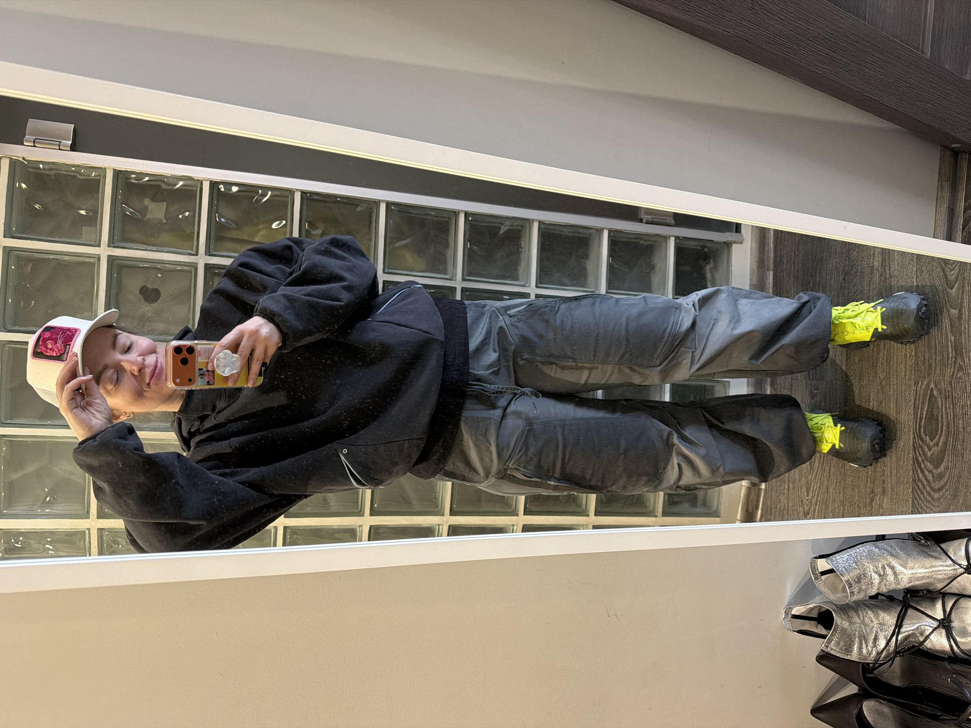 This media file, rotated 90 degrees clockwise, captures a person taking a mirror selfie indoors in Moscow, Russia. The individual is seen from just above the head to their feet, reflected in a large mirror.
The subject is wearing a casual outfit consisting of a black hooded sweatshirt, dark grey or charcoal-colored cargo-style pants, and black athletic shoes with prominent neon yellow accents on the laces and sides. They also wear a white baseball cap, which features a distinct pink rectangular patch on the front, possibly containing the text "VERY RARE" or a similar phrase, though the text is not fully clear. The person holds a smartphone with an orange protective case and a white pop socket attached to the back, using it to take the photograph. They have a relaxed expression, with a slight smile or smirk and their head tilted. Their other hand is resting near their cap.
The background, visible through the mirror's reflection, includes a wall constructed from square glass blocks, suggesting a possibly industrial or older commercial building style. Above the glass blocks, there is a dark grey wall section topped with a light-colored trim and a white ceiling. In the unreflected foreground, beneath the mirror, a white surface holds a stack of shoes, including a pair of shiny silver ankle boots with black laces and some dark-colored footwear.
The overall scene suggests a casual moment, likely in a private or semi-private setting. The lighting is bright and even, consistent with indoor illumination.