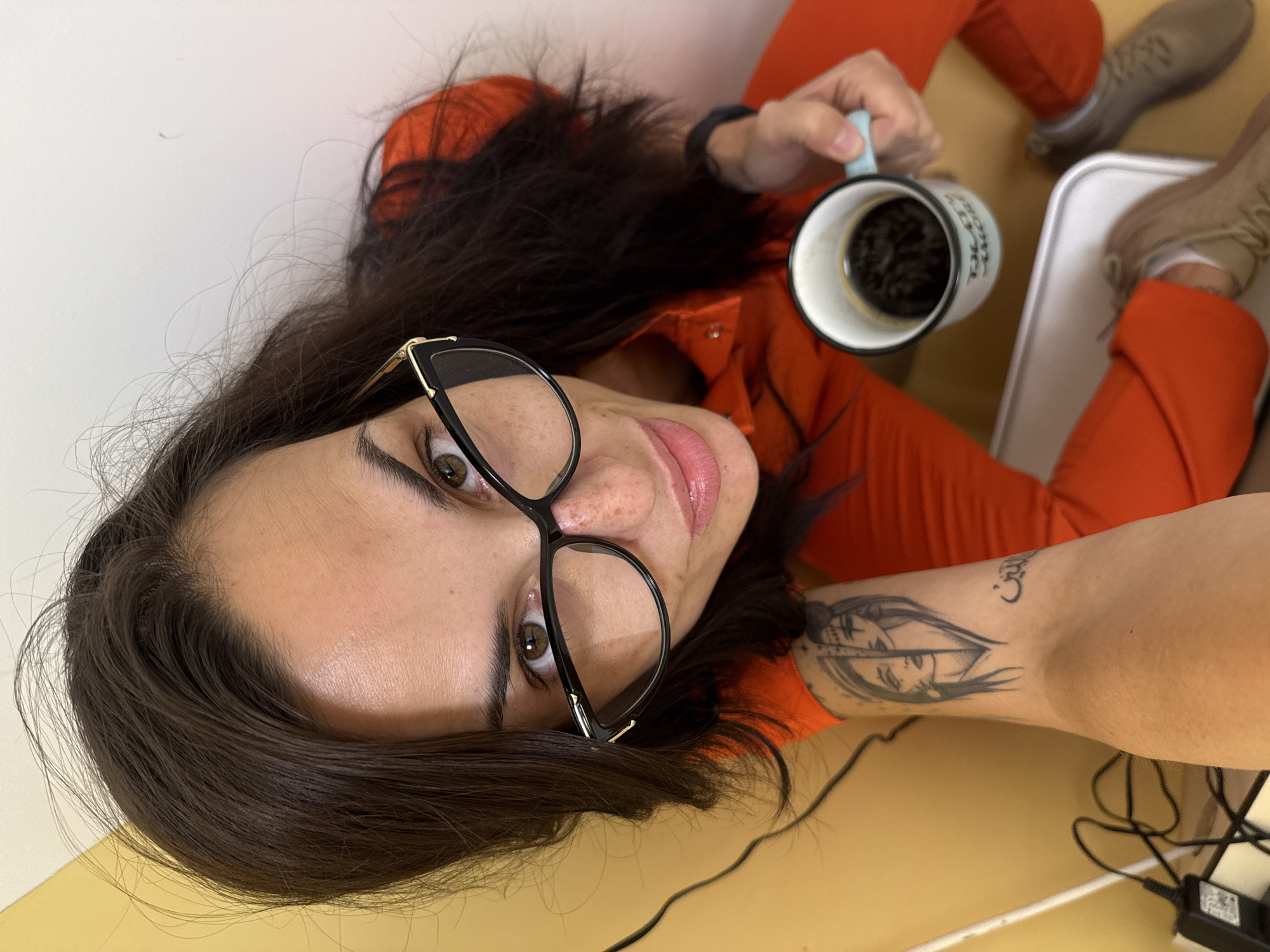 A woman with dark hair and glasses, wearing an orange jumpsuit, is seated on a mustard-colored floor with her legs extended towards a white wall. She is holding a mug of black coffee. Her right arm is visible, featuring a tattoo. Her legs and footwear are in the upper right corner, situated near a white tray. An assortment of wires and an electronic device are present in the lower right corner. The location cannot be identified as Pleven, Bulgaria from the provided information.