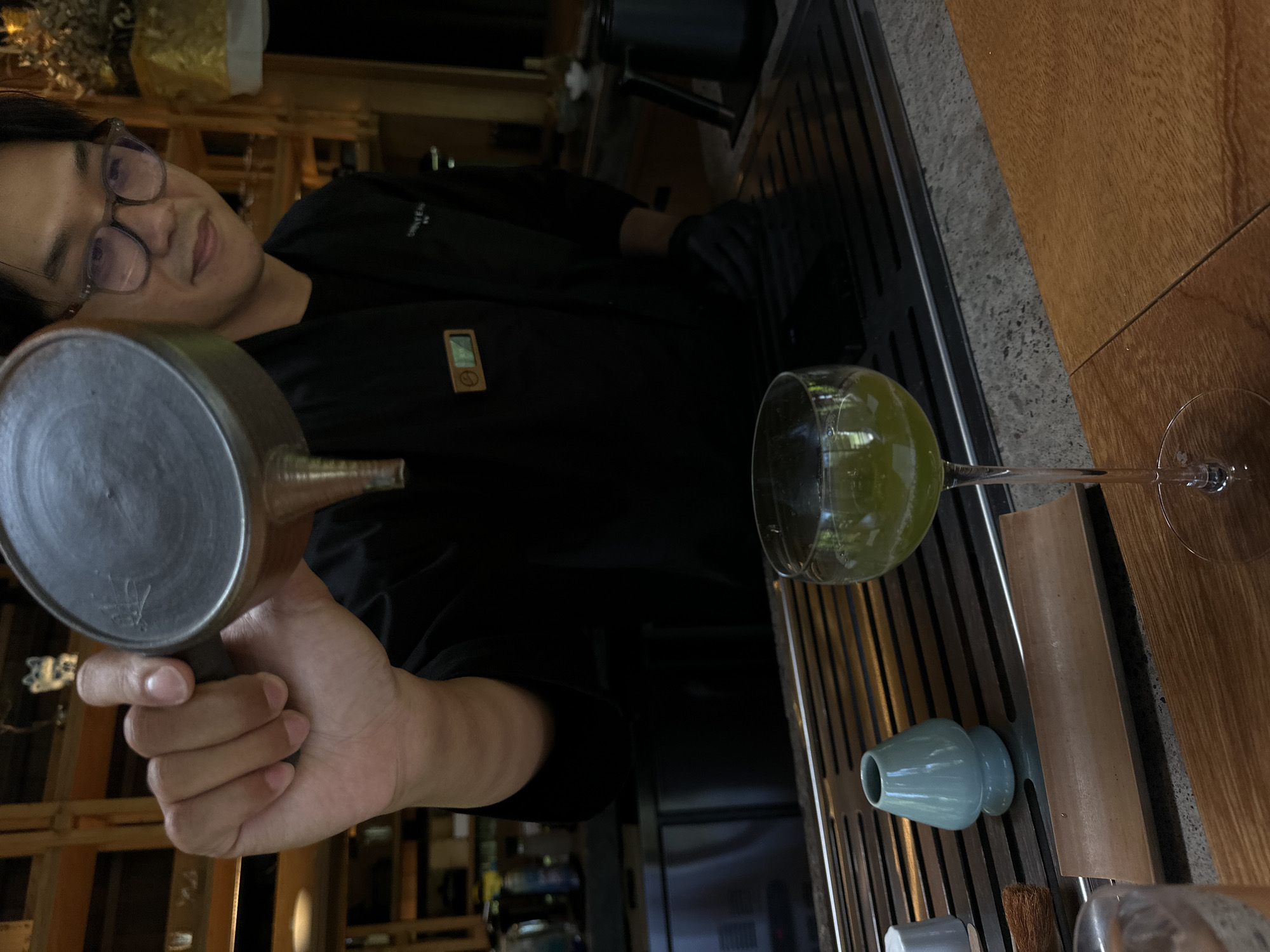 An individual with glasses, possibly a mixologist, is pouring a liquid from a metal vessel into a large wine glass filled with a yellowish-green liquid. The glass is on a metal surface next to a blue ceramic vessel and a small brush. The location is likely a bar or restaurant. Based on the objects and the general style, the scene is located in Ubud, Indonesia.