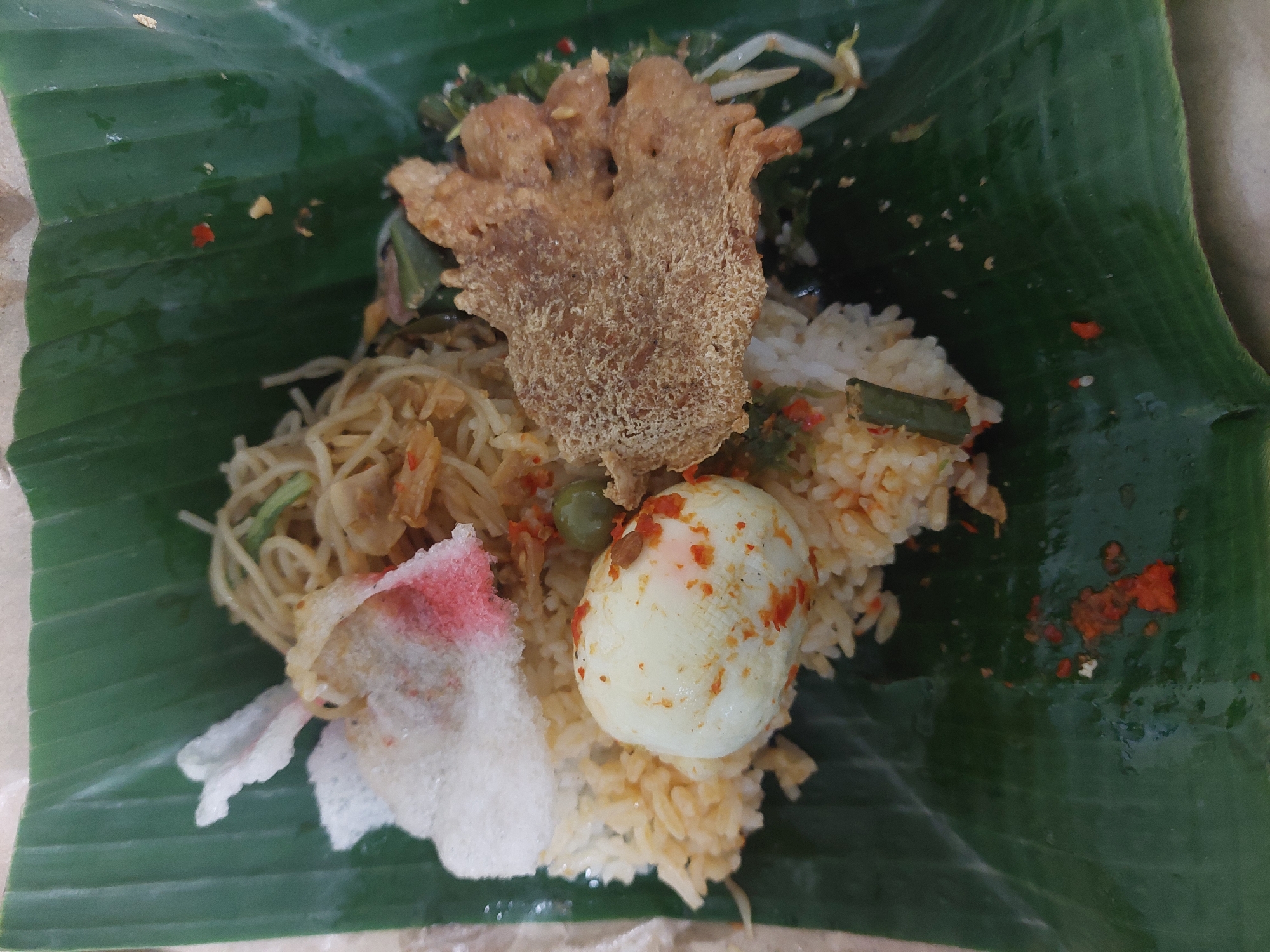 A close-up, overhead shot shows a traditional Indonesian meal served on a banana leaf. The dish consists of rice, a hard-boiled egg with chili flakes, noodles, bean sprouts, and a crispy piece of fried food. The banana leaf is large and green, with visible veins. The meal is arranged in a mound on the leaf, with some chili flakes scattered on the leaf itself. The lighting is natural and soft, highlighting the textures and colors of the food. The background is out of focus, suggesting the photo was taken in a casual dining setting.
