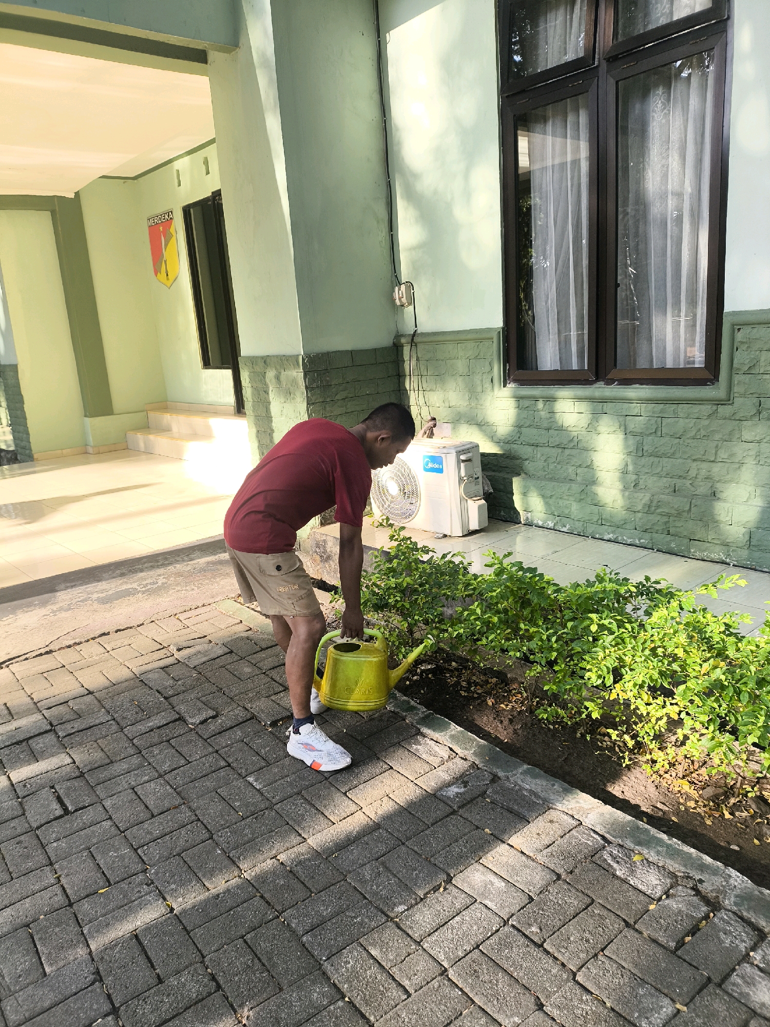 An Indonesian man in a red t-shirt and beige shorts is watering plants with a yellow watering can. He is standing on a paved walkway in front of a light green building with dark brown windows. An air conditioning unit is visible on the side of the building. The building has a yellow and red emblem on the wall. The scene appears to be outdoors, possibly in a courtyard or garden area.