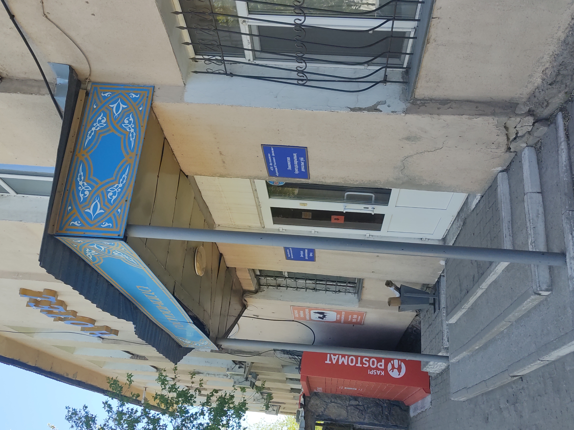 Here is a description of the image:
The image shows a street scene in an urban area. The architecture appears to be a mix of older and more modern styles. In the foreground, there is a "KASPI POSTOMAT" (a post office machine). Above this, there is a building with signage. The most prominent signage reads "PARIKMAHERSKAYA" (hair salon). In addition, a sign in what appears to be Kazakh reads "«бураганди ИТ бөлікшесі филиалы» Локомотив бригадаларының демалыс үйі". The blue and gold decorative columns and awning suggest a business or institution with a sense of civic pride. The building appears to be located on a relatively quiet street with trees nearby.