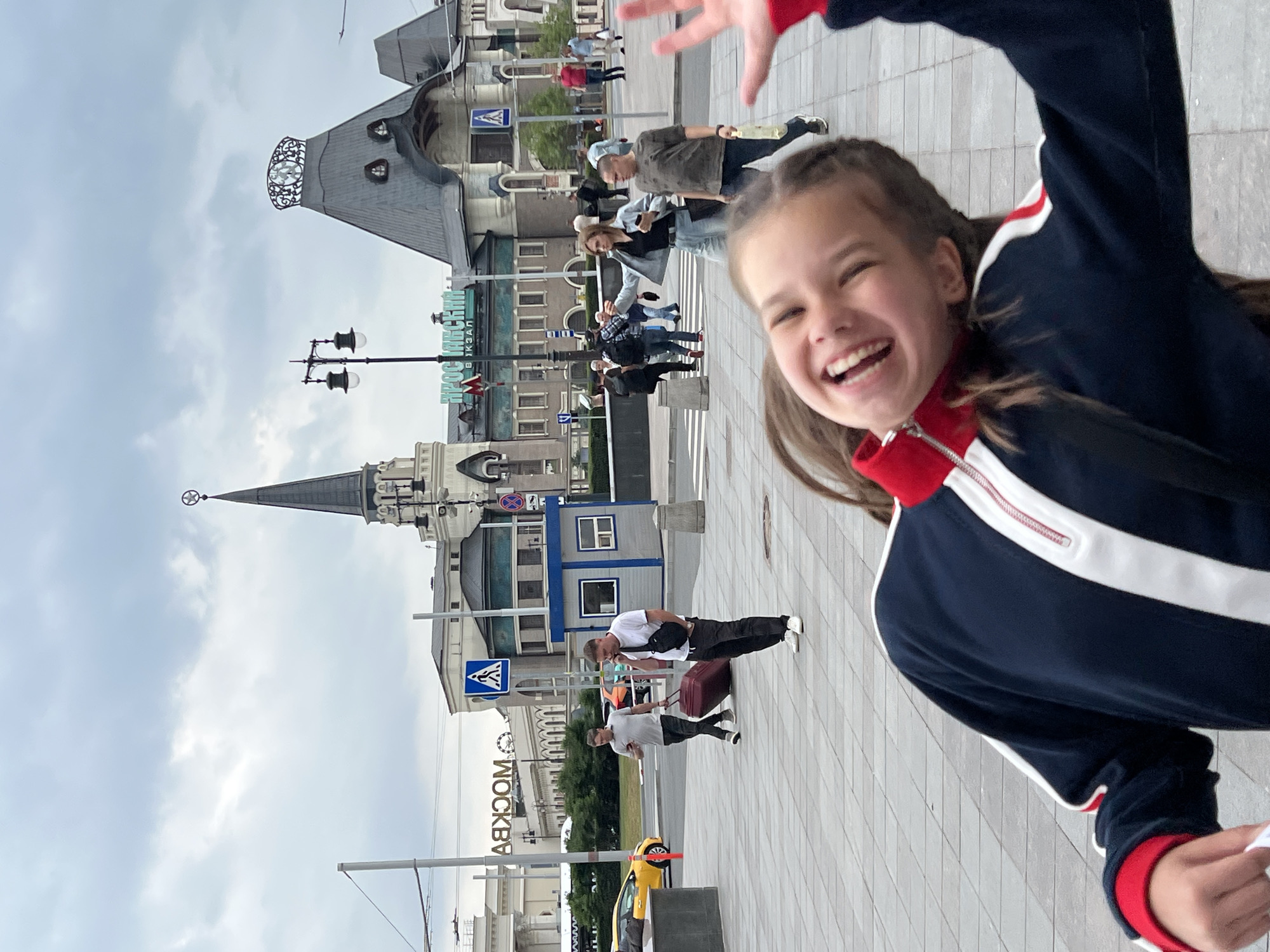 The image captures a lively scene outside a train station in Moscow, Russia, likely Komsomolskaya Square, given the iconic architecture. In the foreground, a young girl with pigtails is the primary subject, her face contorted in a wide, joyful smile, her mouth open as she laughs. She is wearing a dark blue and white zip-up jacket with red trim. Her right arm is raised, and her hand is positioned in a wave, suggesting interaction with the photographer.
The background features a bustling environment with several people. A man in a white t-shirt and dark pants is talking on his phone while holding a black bag across his chest, standing near a small blue and grey booth. Another man is pulling a large, dark red suitcase. Behind them, a yellow car is visible on the street, and further back, the distinctive architecture of the train station dominates the skyline, with a tall, pointed tower topped with a star and a building with a unique, dark grey, pitched roof. Numerous other individuals are scattered across the paved area, some walking, others standing, contributing to the sense of activity. The sky is overcast with a pale grey hue. The ground is paved with large, light grey tiles.