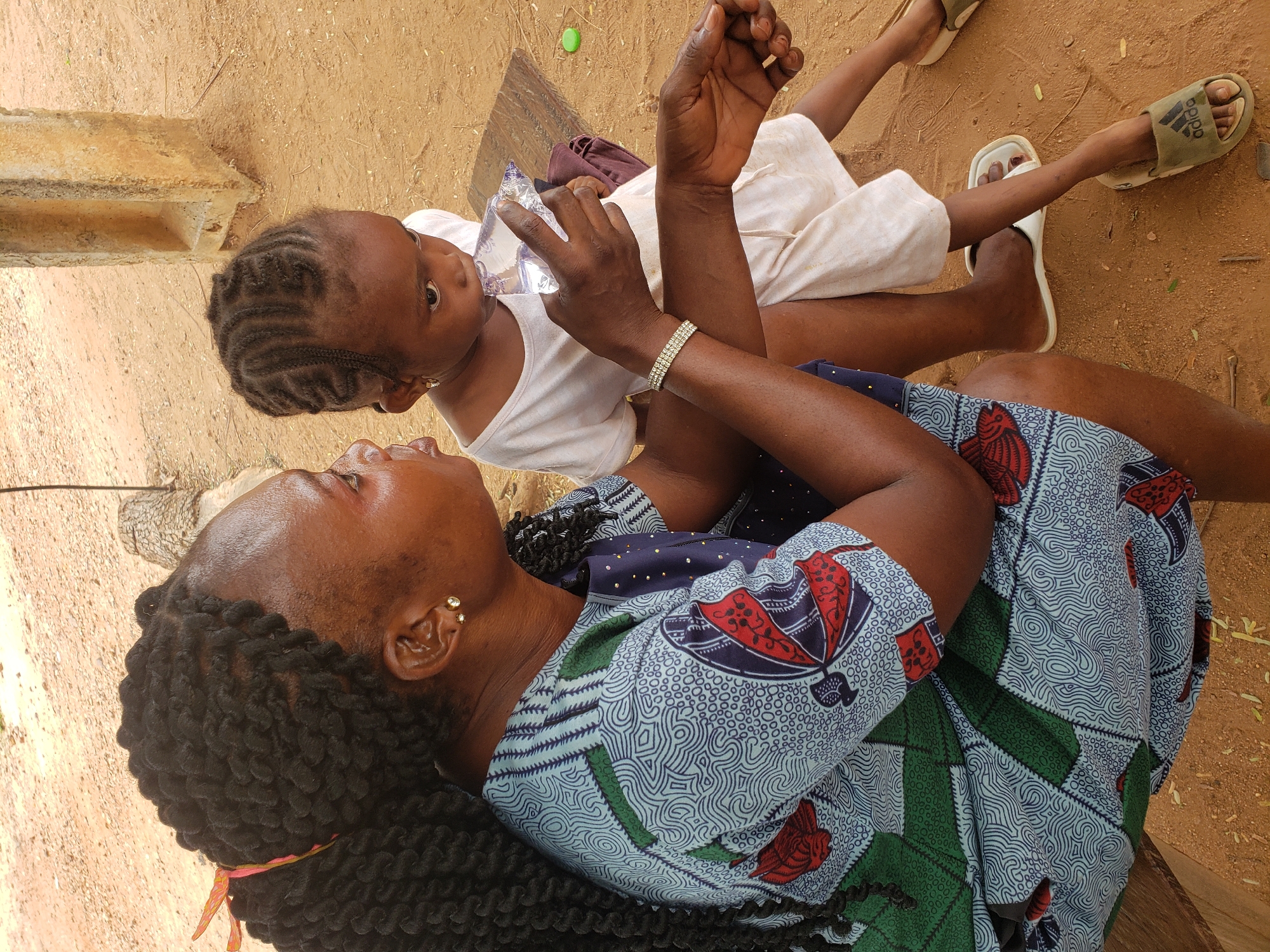 An overhead shot captures two females, an adult and a young child, interacting on a dirt ground in Adankolo, Nigeria, under bright daylight. The adult woman lies on her back, looking up towards the child, who is seated beside her.
The adult has dark skin and black, twisted braids. She wears multiple small gold stud earrings and a two-row rhinestone bracelet on her right wrist. Her attire is a vibrant, patterned dress featuring geometric and floral designs in blue, red, green, and white. Her right hand is holding a clear plastic water bottle, seemingly offered to the child.
The young child, also with dark skin, has black hair styled in neat cornrows and a small gold earring. They are dressed in a plain white t-shirt and light-colored shorts. One foot is bare, while the other wears a green slide sandal with the "adidas" logo visible on the strap.
The setting is an outdoor, earthy environment with light brown dirt ground and a few scattered dry leaves. A simple, light-colored concrete block is visible in the background. The scene portrays a tender moment of care and connection between the two individuals.