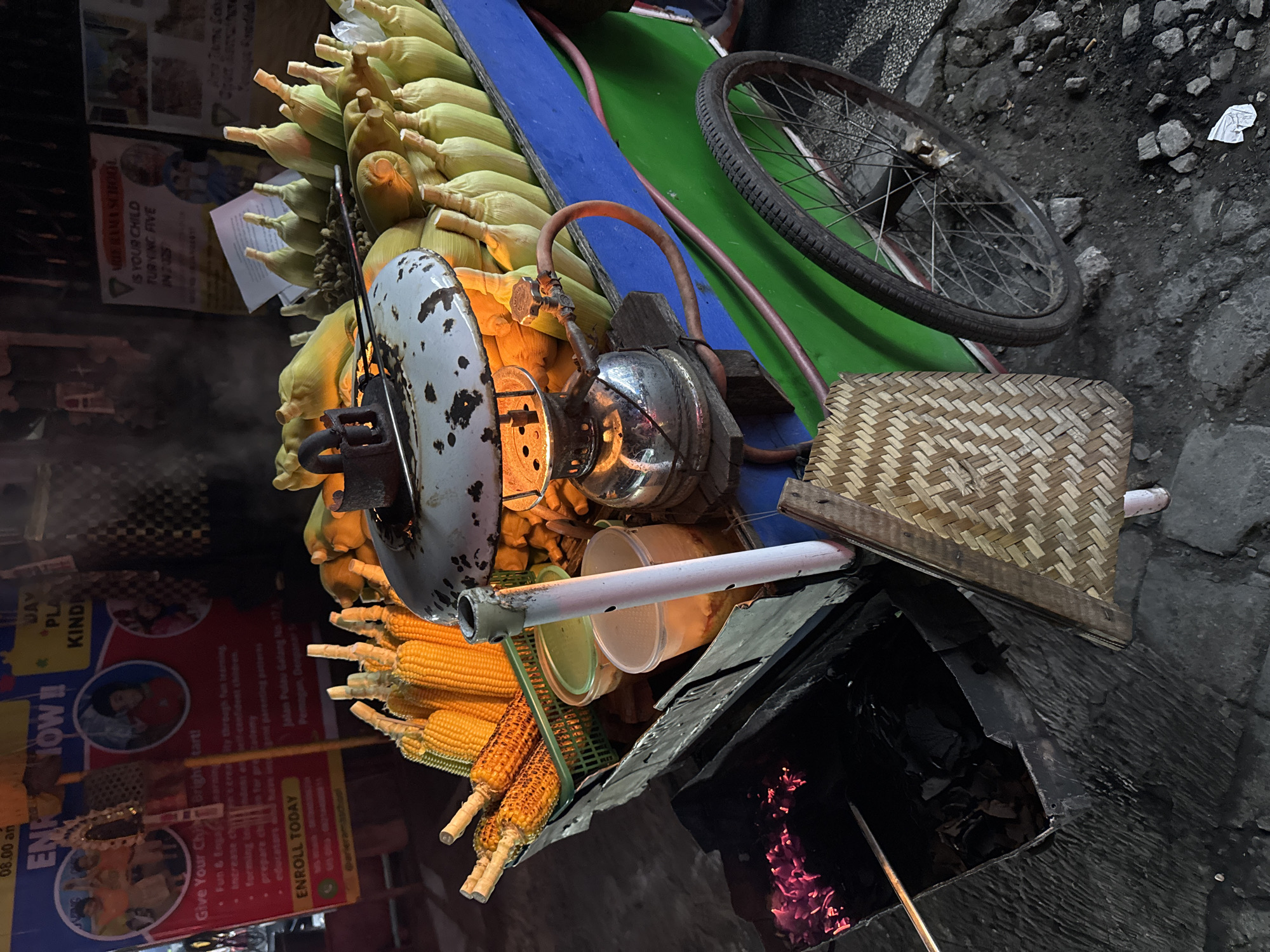 The image depicts a street food cart on a paved surface in Denpasar, Indonesia. The cart is laden with numerous ears of corn, some with husks still attached and others roasted to a golden brown. A gas-powered lamp, likely used for roasting or keeping the corn warm, is prominently featured on the cart, emitting a warm glow. To the left of the cart, a charcoal grill with glowing embers suggests that some of the corn is being roasted over an open flame. A green basket holds some of the roasted corn. Various plastic containers, possibly for sauces or toppings, are also visible. The cart has a green and blue painted body and is equipped with a spoked wheel, indicating it is mobile. In the background, posters advertising a school are displayed, suggesting a location near an educational institution. The overall scene captures the ambiance of a bustling night market or street food vendor in Denpasar.