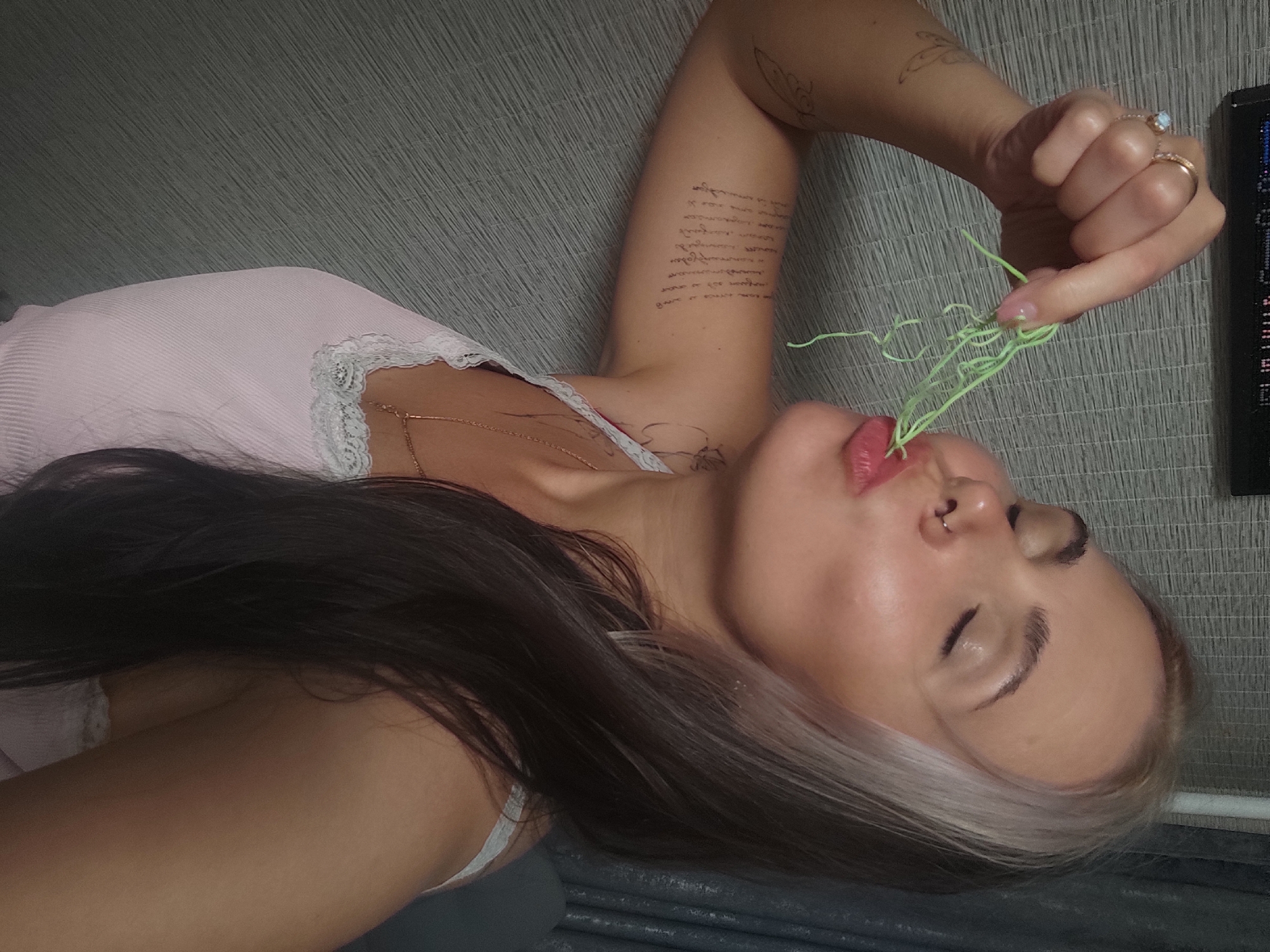 The image features a female subject in a mid-shot, positioned against a textured indoor wall. She is shown with her eyes closed and lips pursed, holding a bundle of thin, light green, string-like material to her mouth with her right hand.
The subject has long, dark brown hair with a prominent light blonde or silver streak framing the left side of her face. A gold-colored septum ring is visible in her nose. Her right hand displays two rings: a band with a light-colored, possibly iridescent stone on her ring finger, and a plain band on her middle finger. Her fingernails are painted a light pink.
She is wearing a light pink, ribbed tank top with white lace trim along the neckline. A thin, gold-colored chain necklace is visible around her neck. Tattoos are present on her arms; a significant section of script or text runs vertically down her left bicep, and fainter, less distinct outlines are visible near her left shoulder and on her right shoulder area.
The background consists of a light gray or beige wall with a consistent horizontal ribbed or woven texture. In the upper left corner, a dark, rectangular object, possibly an electronic device or shelf, is partially visible. The lighting appears to be artificial, highlighting the subject from the front or side. There are no identifiable features within the image that indicate a specific city or country.