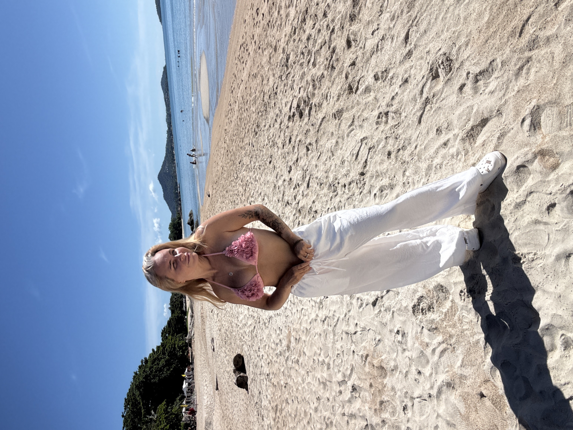 A blonde woman with tattoos on her arms is standing on a sandy beach in Baan Chaweng Noi, Thailand. She is wearing a pink floral bikini top and loose white pants. The beach has a light brown sand with some footprints and debris. In the background, there are palm trees, some white umbrellas, and the ocean. The water is blue with some gentle waves. There are a few people in the ocean in the distance. The sky is clear blue with a few wispy clouds. The sun is shining brightly, casting shadows on the sand.