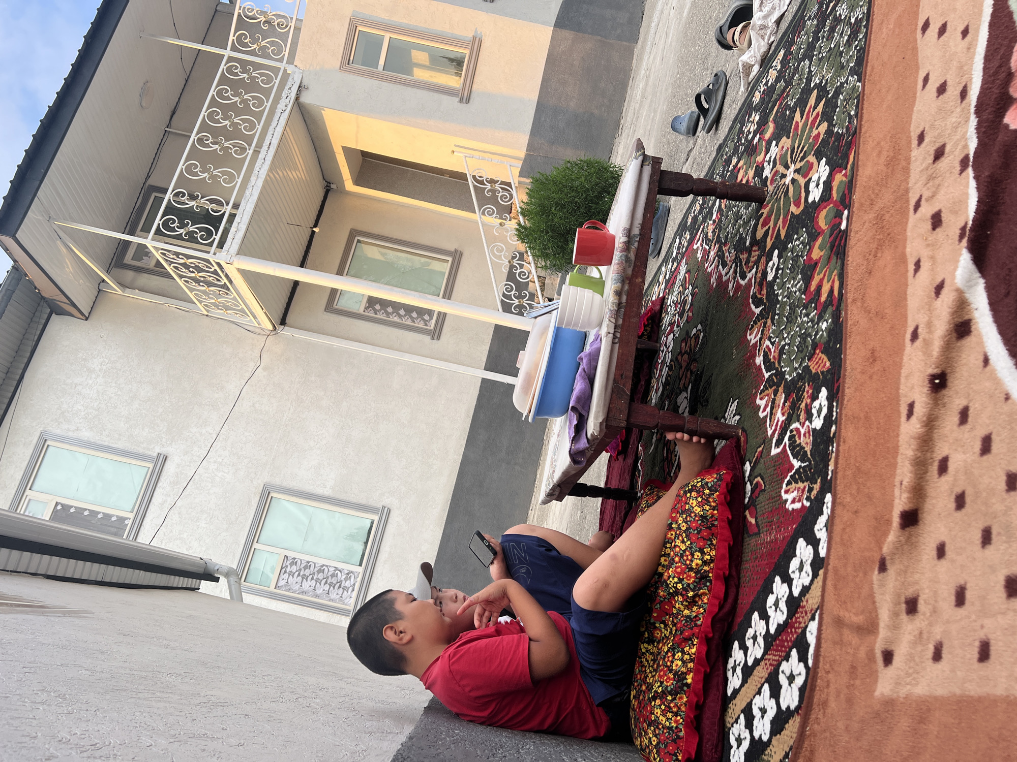 In Kara-Dıykan, Kyrgyzstan, two boys are seated on the ground next to a wall, engaged with a mobile phone. The elder boy, wearing a red t-shirt and blue shorts, holds the phone and appears to be showing it to the younger boy, who is wearing a grey cap and a black shirt. They are positioned on a large, patterned cushion with floral designs.
In front of them, a low wooden table, covered with a patterned cloth, holds several items. A blue plastic container, two mugs (one red, one green), and a stack of white bowls are visible on the table. A purple cloth is draped over the edge of the table. The ground is covered with a large, intricately patterned rug, extending out from under the table and cushion.
Behind the boys and the table, a two-story building stands. The lower part of the building is a light grey, while the upper portion is a lighter, off-white color. The building features several windows with decorative grey frames. A white-railed balcony extends from the upper floor, supported by white columns. The scene is illuminated by the warm light of the setting sun, casting long shadows and highlighting the textures of the building and the surrounding area. The ground appears to be a paved surface, possibly concrete, with some scattered footwear visible. A dense green shrub is planted near the building, between the table and the steps leading to the balcony.