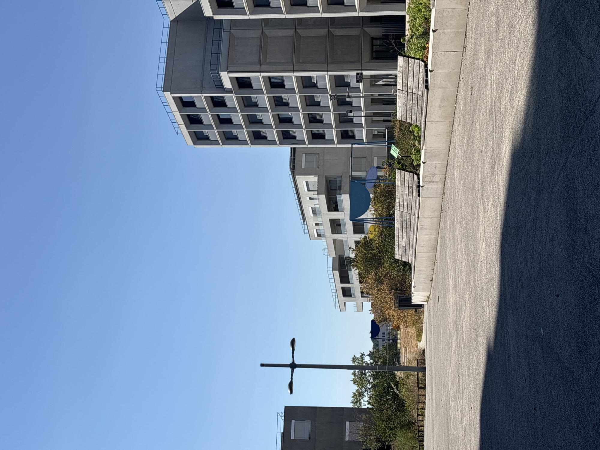 The image captures a modern urban landscape under a clear blue sky, likely in Lyon, France. The scene is dominated by several large, concrete residential buildings with a minimalist architectural style, characterized by repetitive window patterns and angular facades. In the foreground, a paved area, possibly a plaza or walkway, is visible, with two wooden benches situated along a low concrete wall. Beyond the benches and greenery, a lamppost with two lights stands tall. Further in the background, more buildings and trees are discernible, suggesting a developed urban environment. The overall impression is one of contemporary city living with an emphasis on concrete structures and open public spaces.